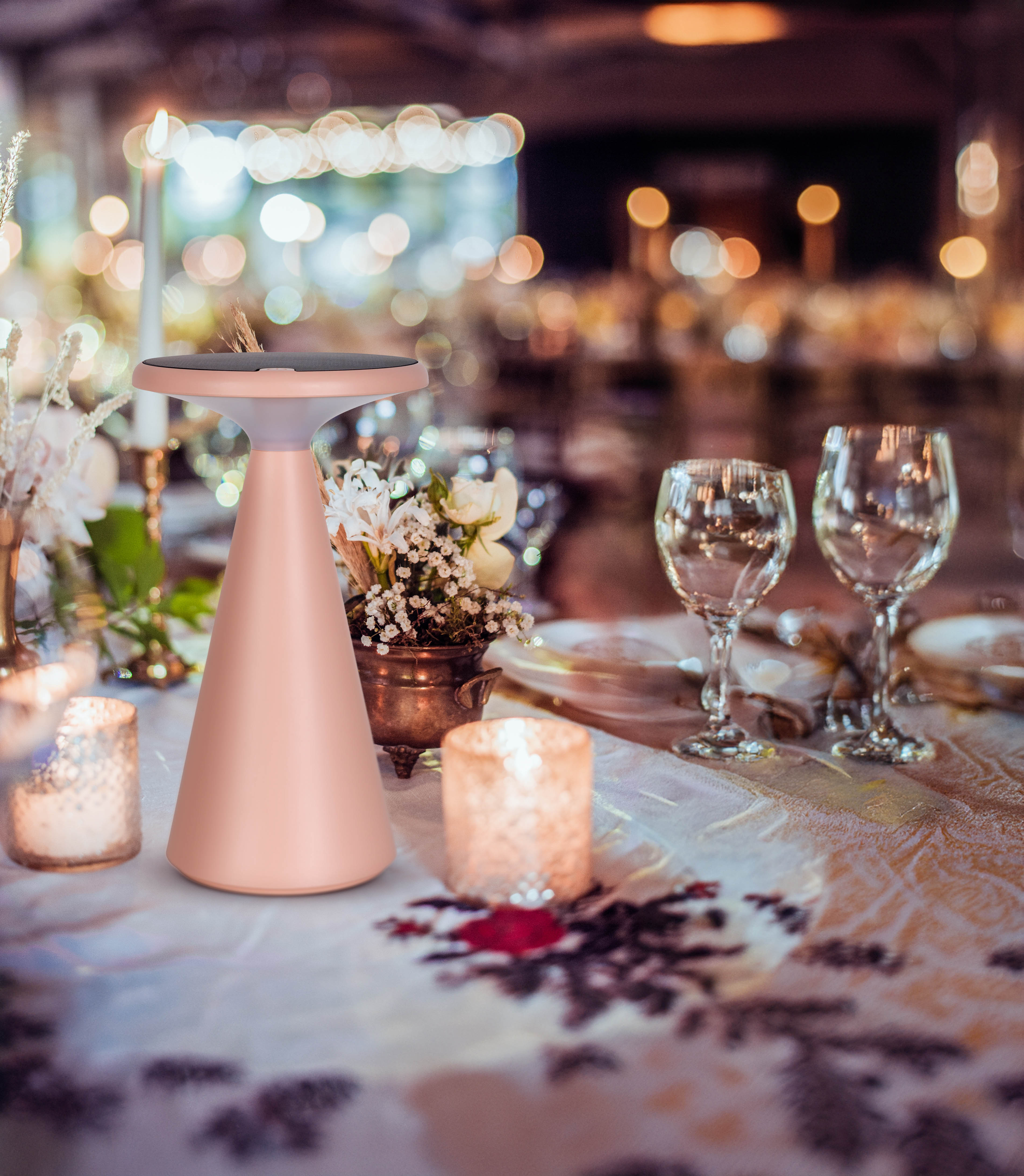 Eine roségoldene Tischlampe auf einem Tisch mit Kerzen und Blumen, im verschwommenen Hintergrund.
