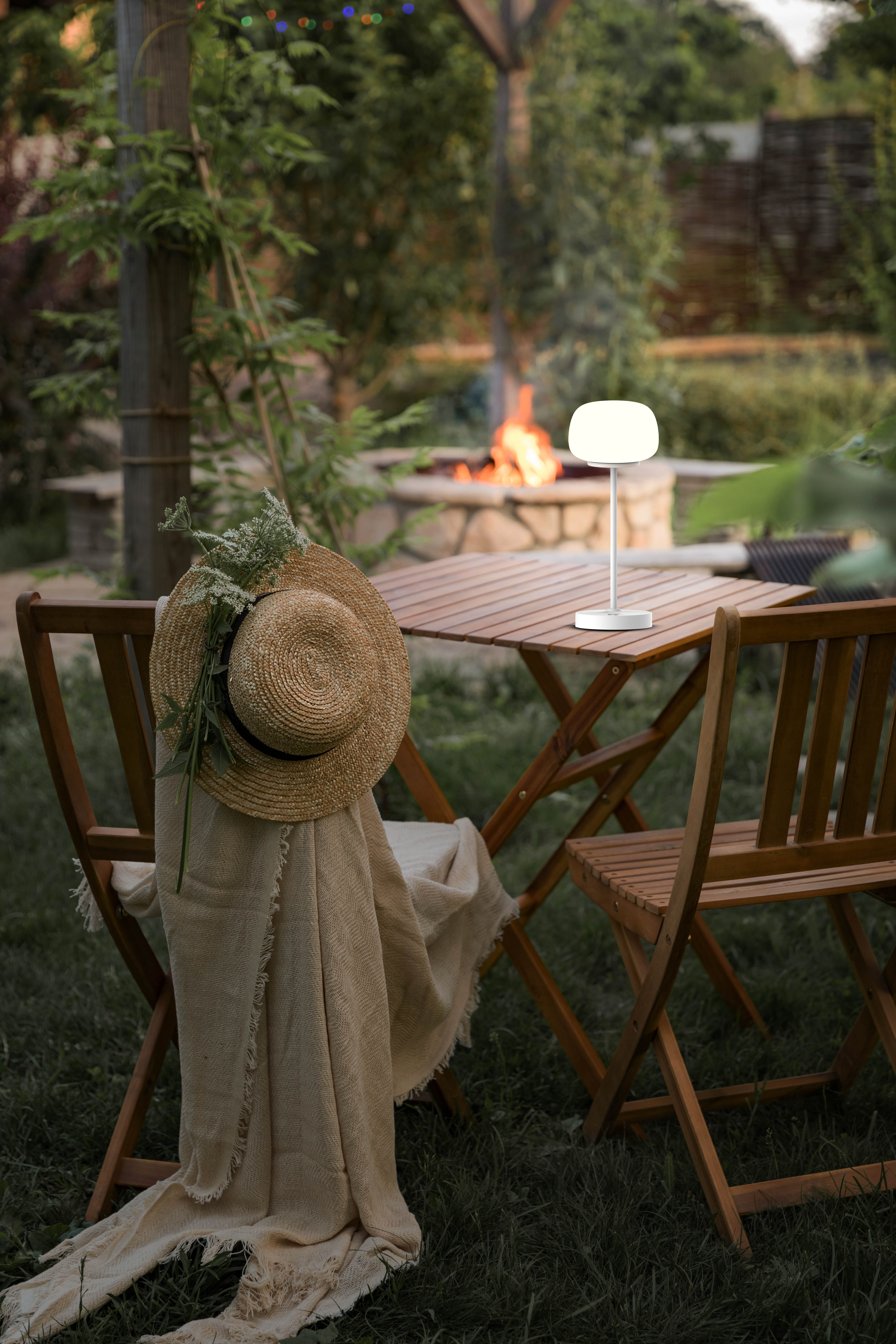 Szene im Freien mit Tisch, Stühlen, Lampe, Hut und Feuerstelle im Hintergrund.