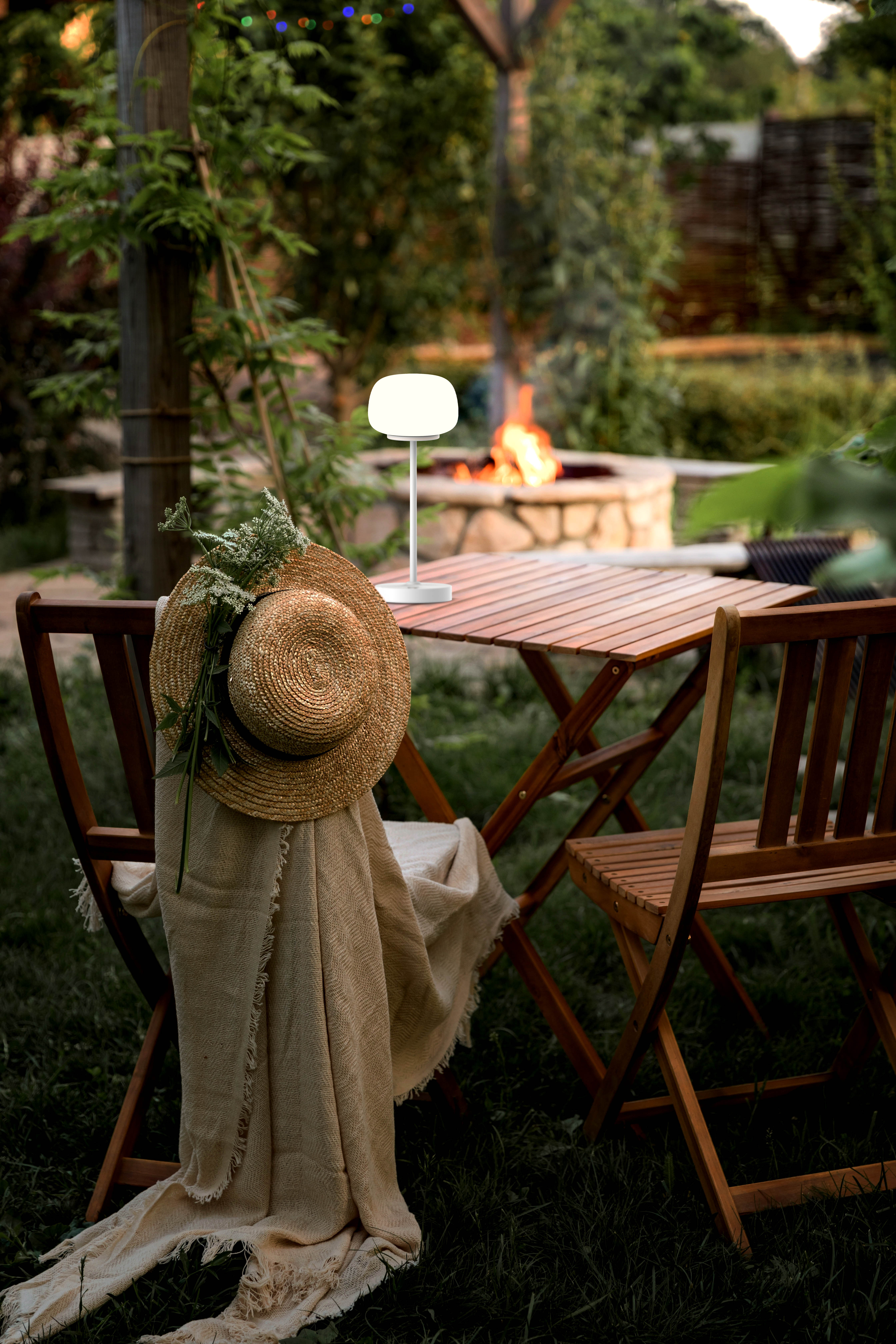Szene im Freien mit Tisch, Stühlen, Hut und Lampe mit Lagerfeuer im Hintergrund.