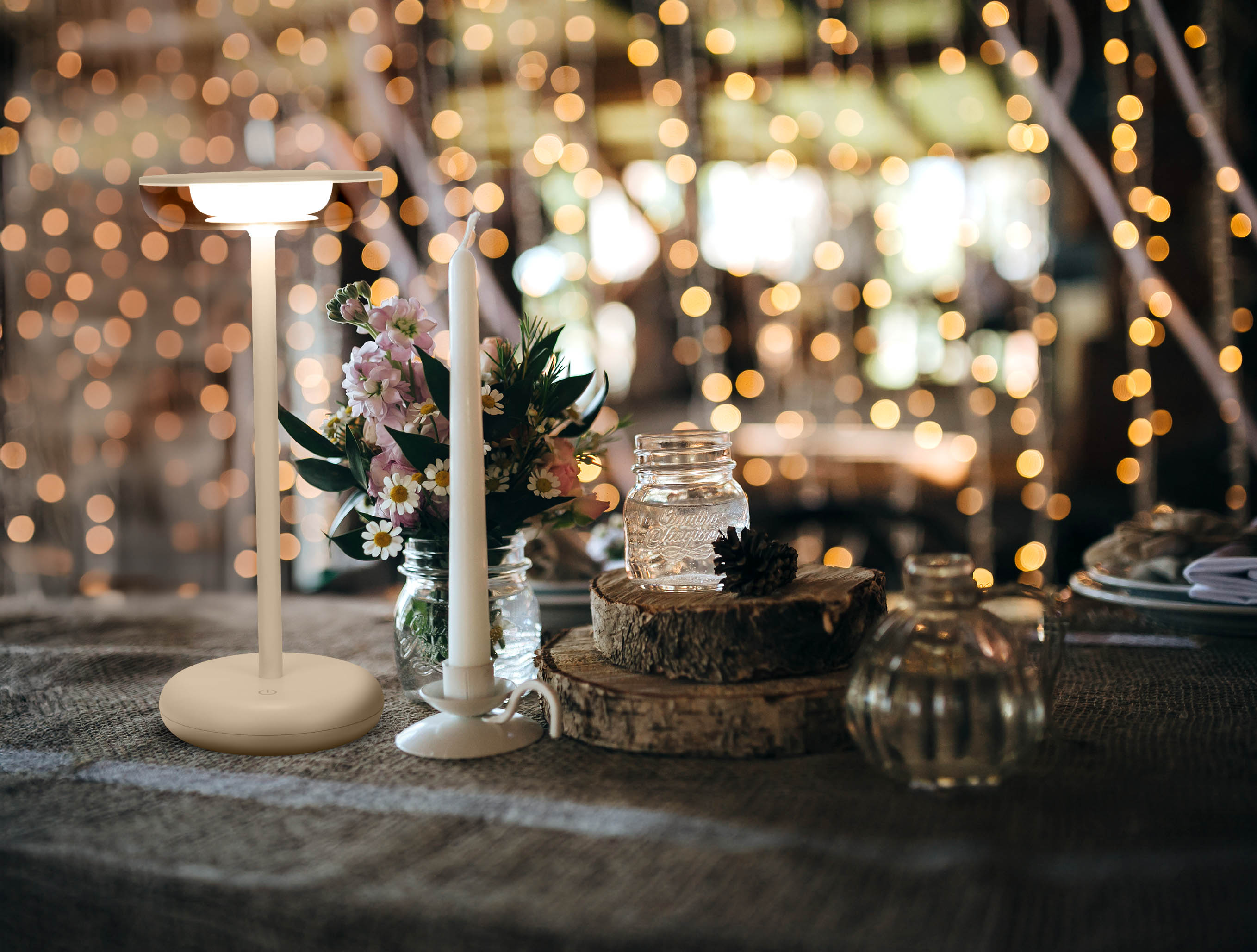 Eine Tischdekoration umfasst eine Lampe, eine Kerze, Blumen und ein Glas. Der Hintergrund hat Lichter.