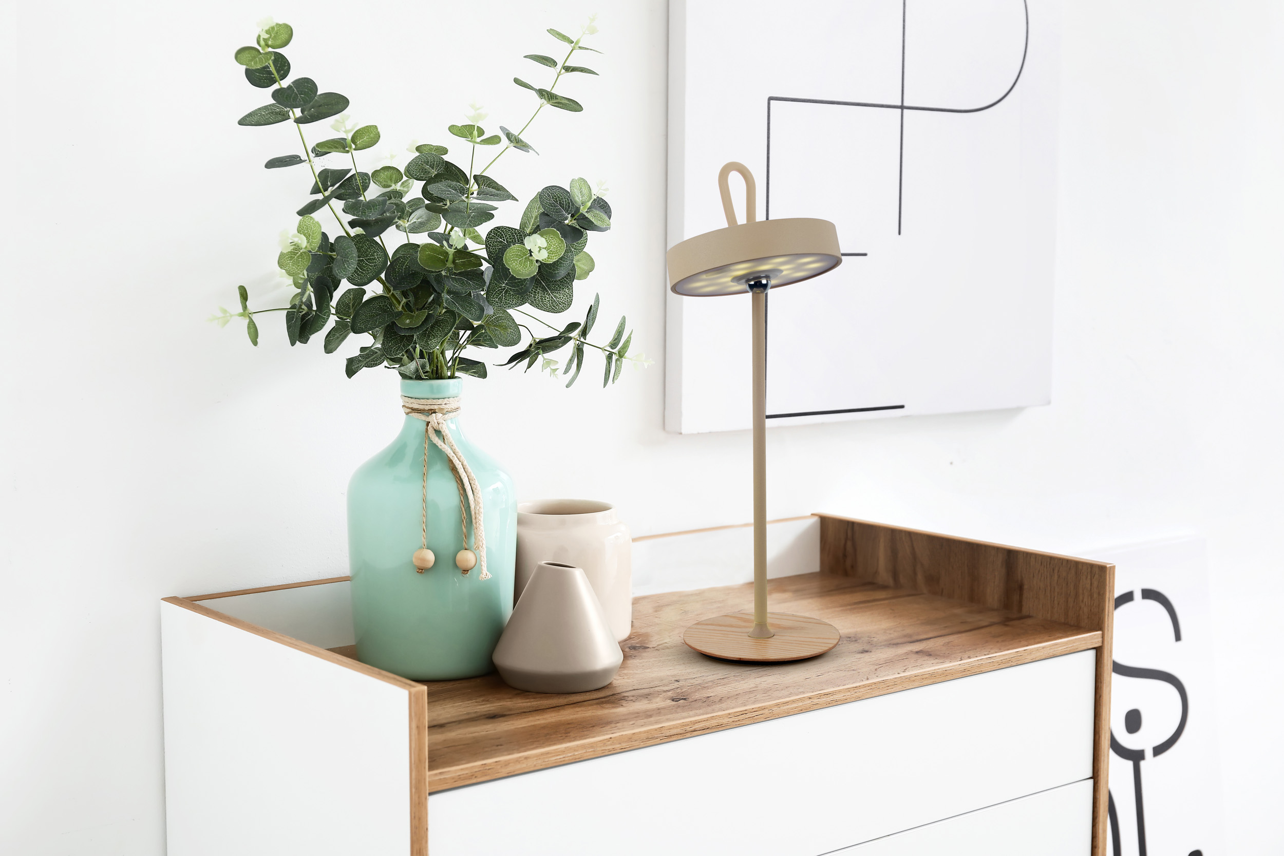 Ein Nachttisch mit Lampe, Vase mit Blättern und Dekoartikeln. Hintergrund: weiße Wand.