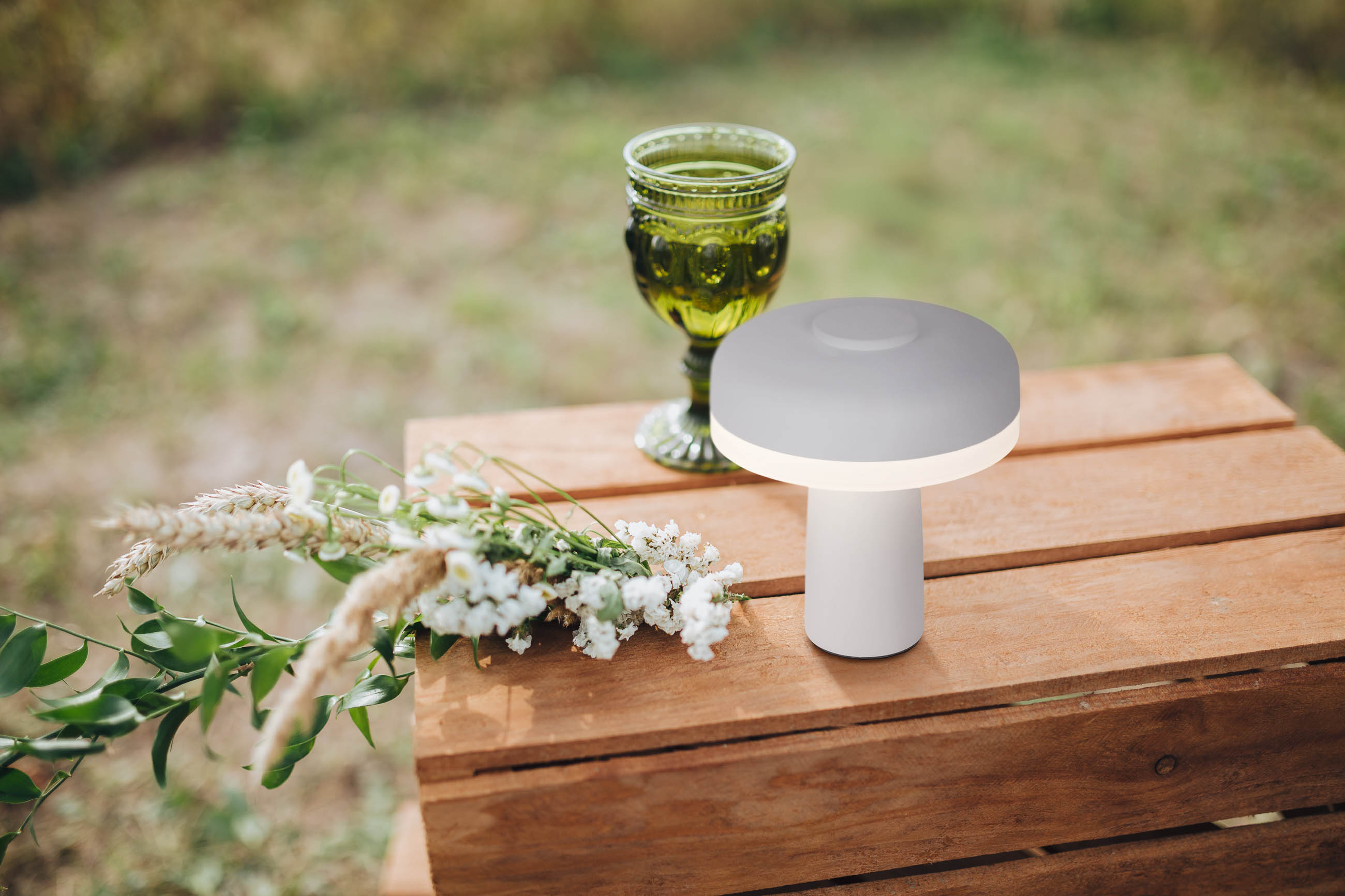 Eine weiße Lampe auf einer Holzkiste, mit einem grünen Glas und Blumen.