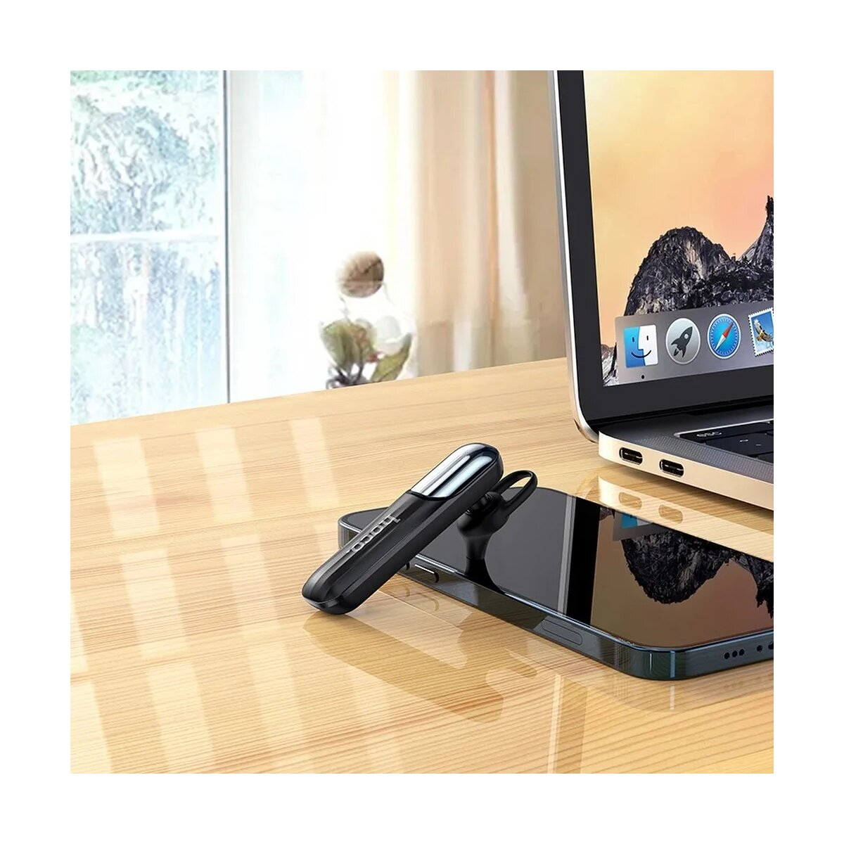 Ein schwarzes Bluetooth-Headset liegt auf einem Smartphone neben einem Laptop auf einem Holztisch. Im Hintergrund ist ein Fenster.
