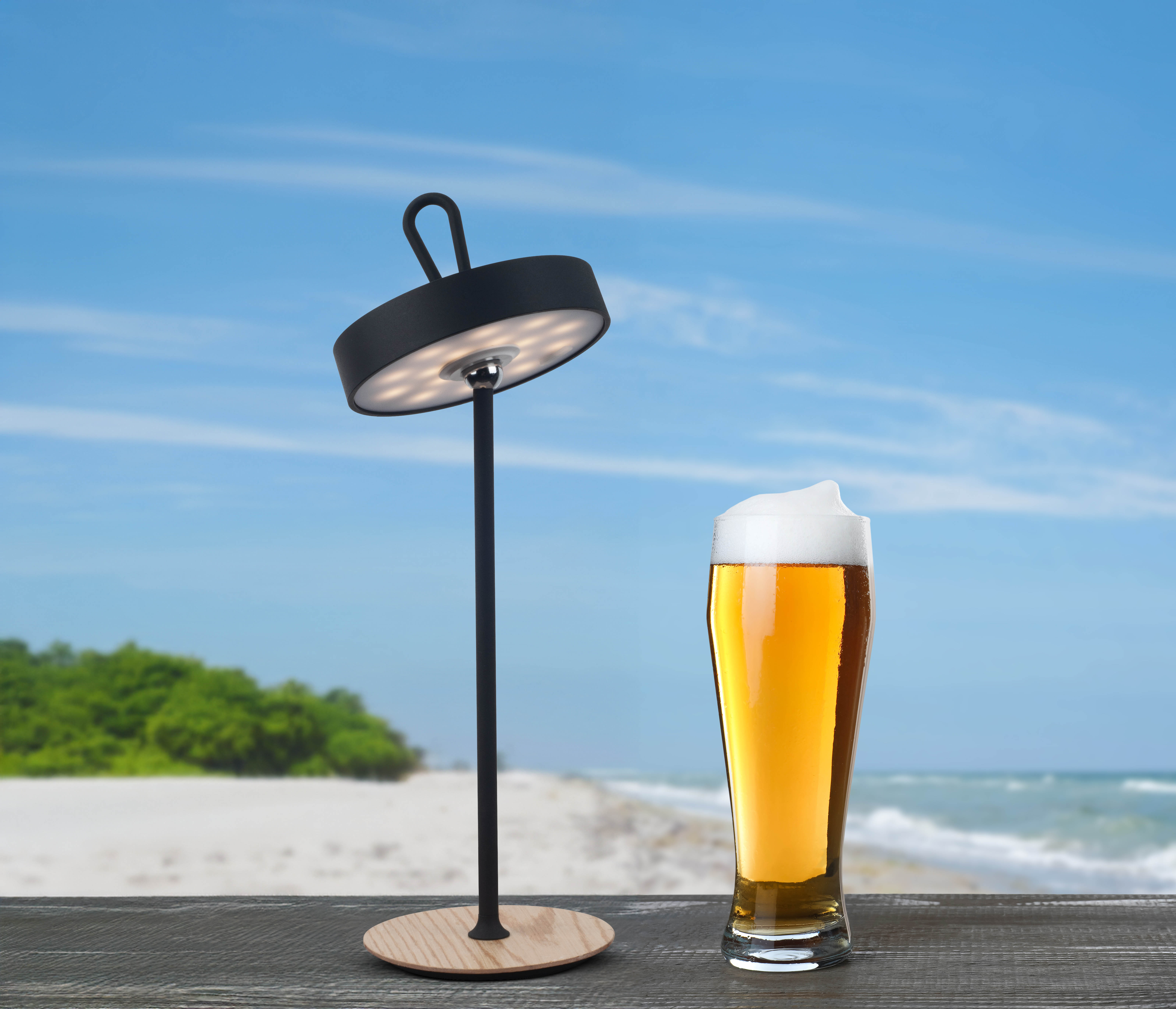 Eine moderne schwarze Lampe und ein Bierglas auf einem Holztisch, Strandhintergrund.