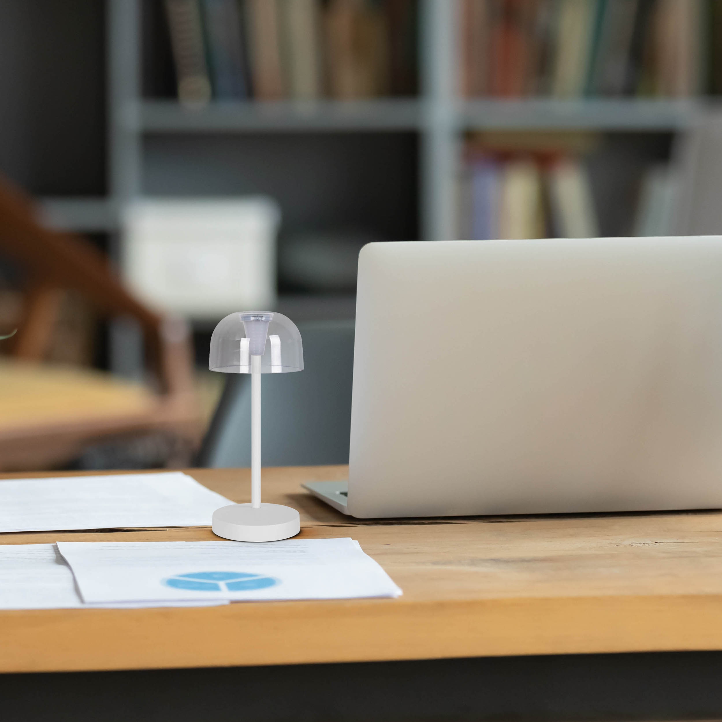 Kleine weiße Schreibtischlampe auf einem Holztisch mit Laptop und Papieren im Hintergrund.
