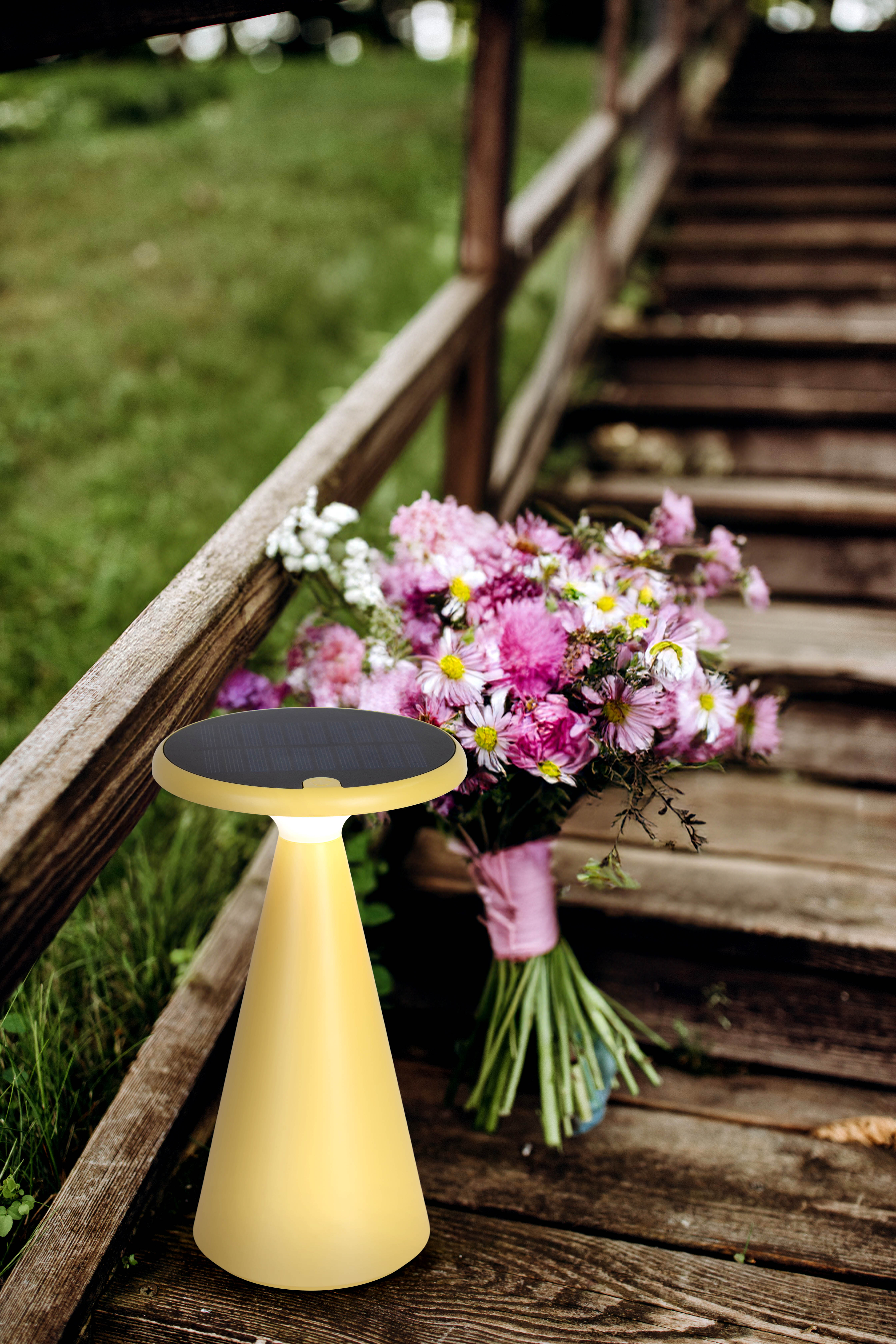 Eine gelbe Lampe steht auf Holzstufen, neben einem Blumenstrauß. Holzgeländer und grünes Gras im Hintergrund.