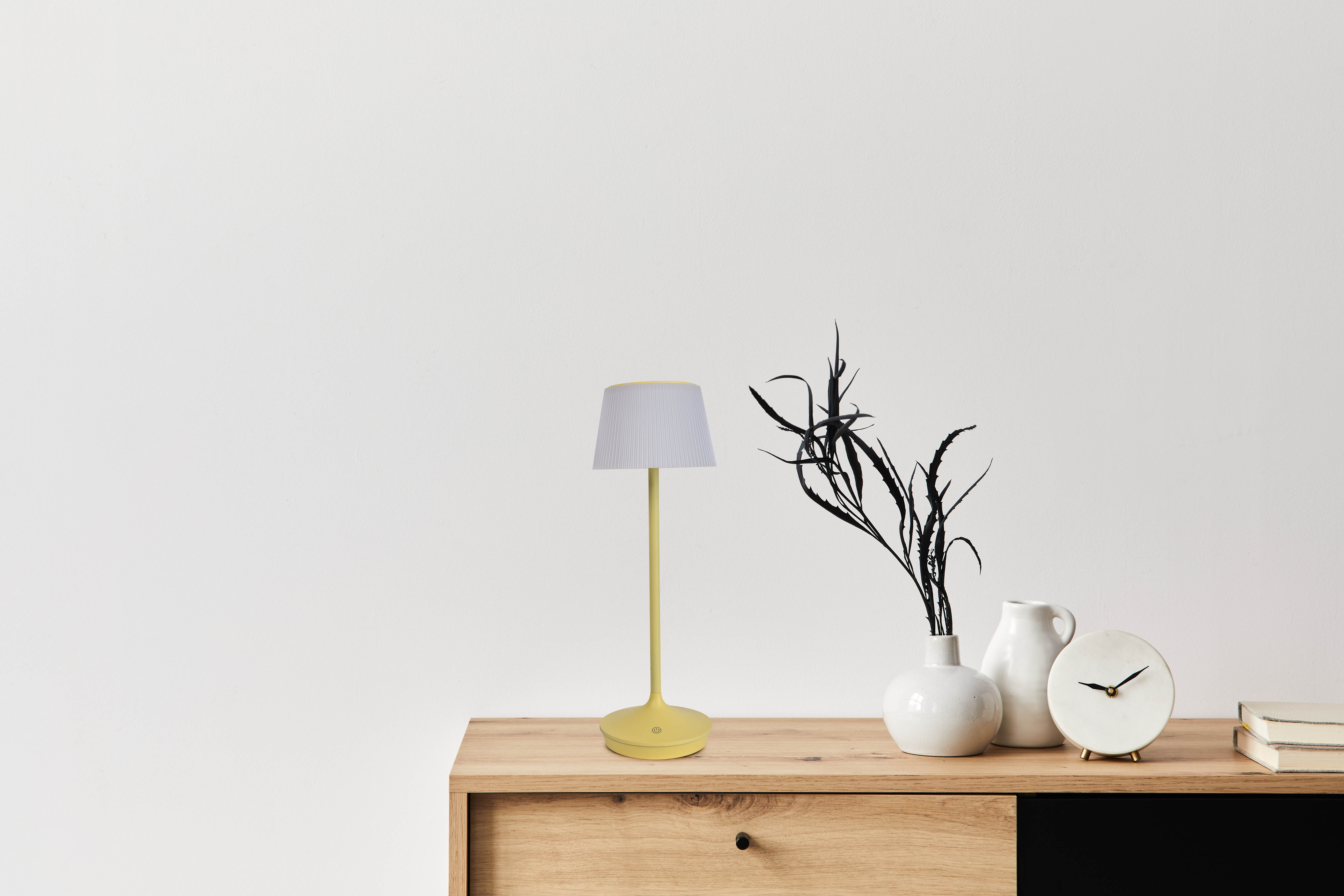 Eine gelbe Lampe mit weißem Lampenschirm und ein Holztisch mit weißen Vasen und einer Uhr.