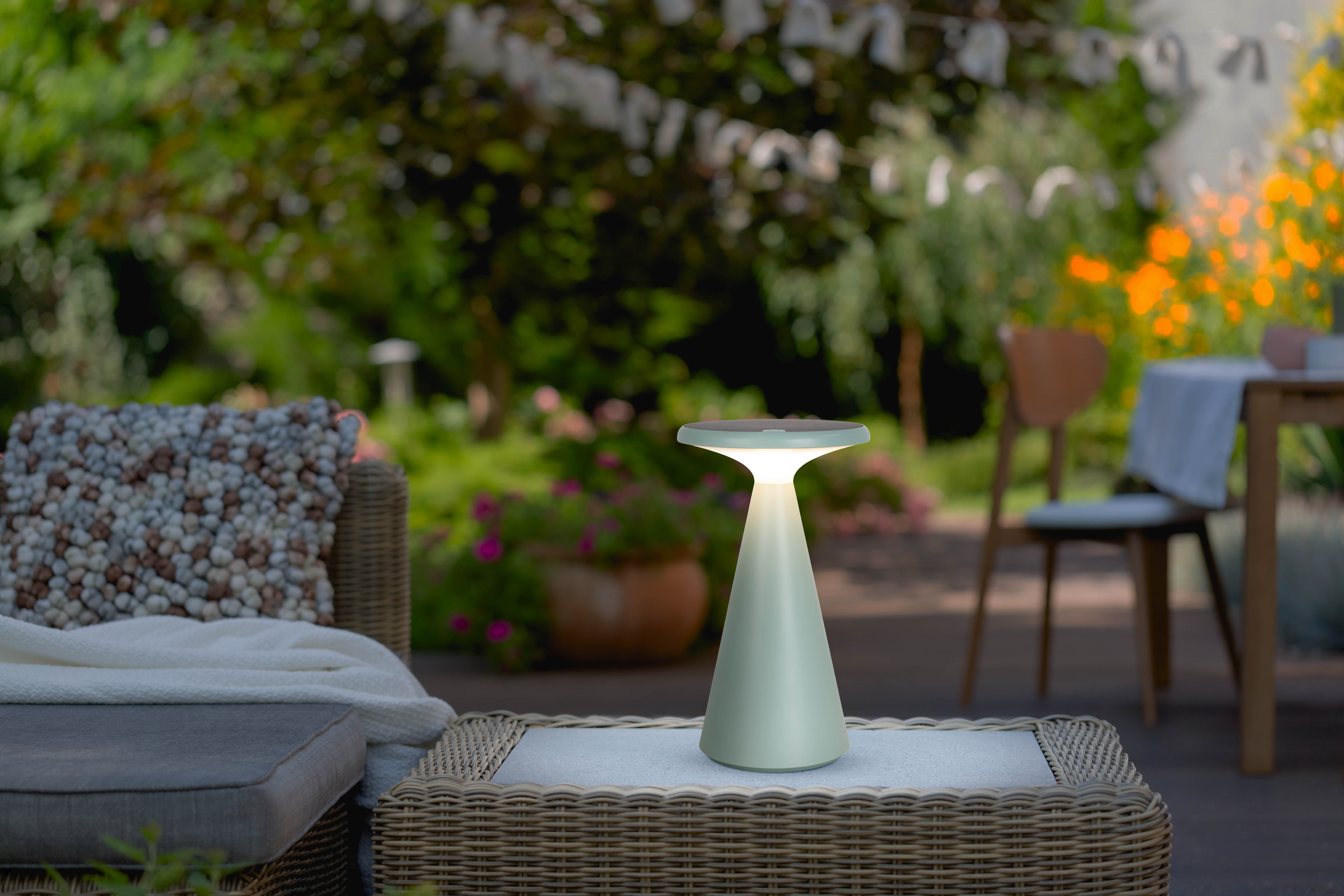 Eine weiße und grüne Außenlampe auf einem Tisch. Der Hintergrund ist ein Garten mit Bäumen und Pflanzen.