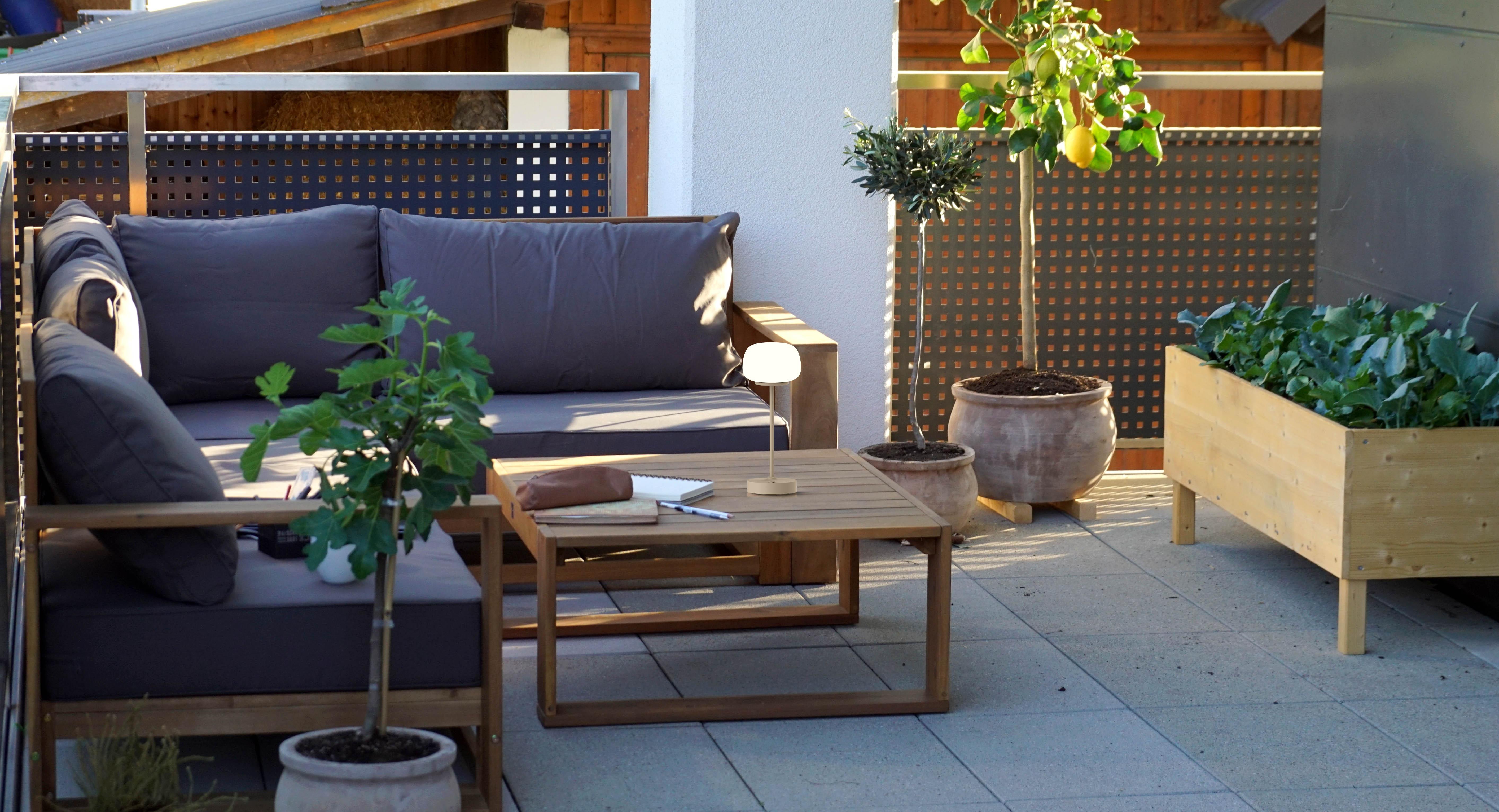 Balkonszene: graues Sofa, Holztisch, Pflanzen und eine Lampe. Im Hintergrund ein Zaun und Pflanzen.