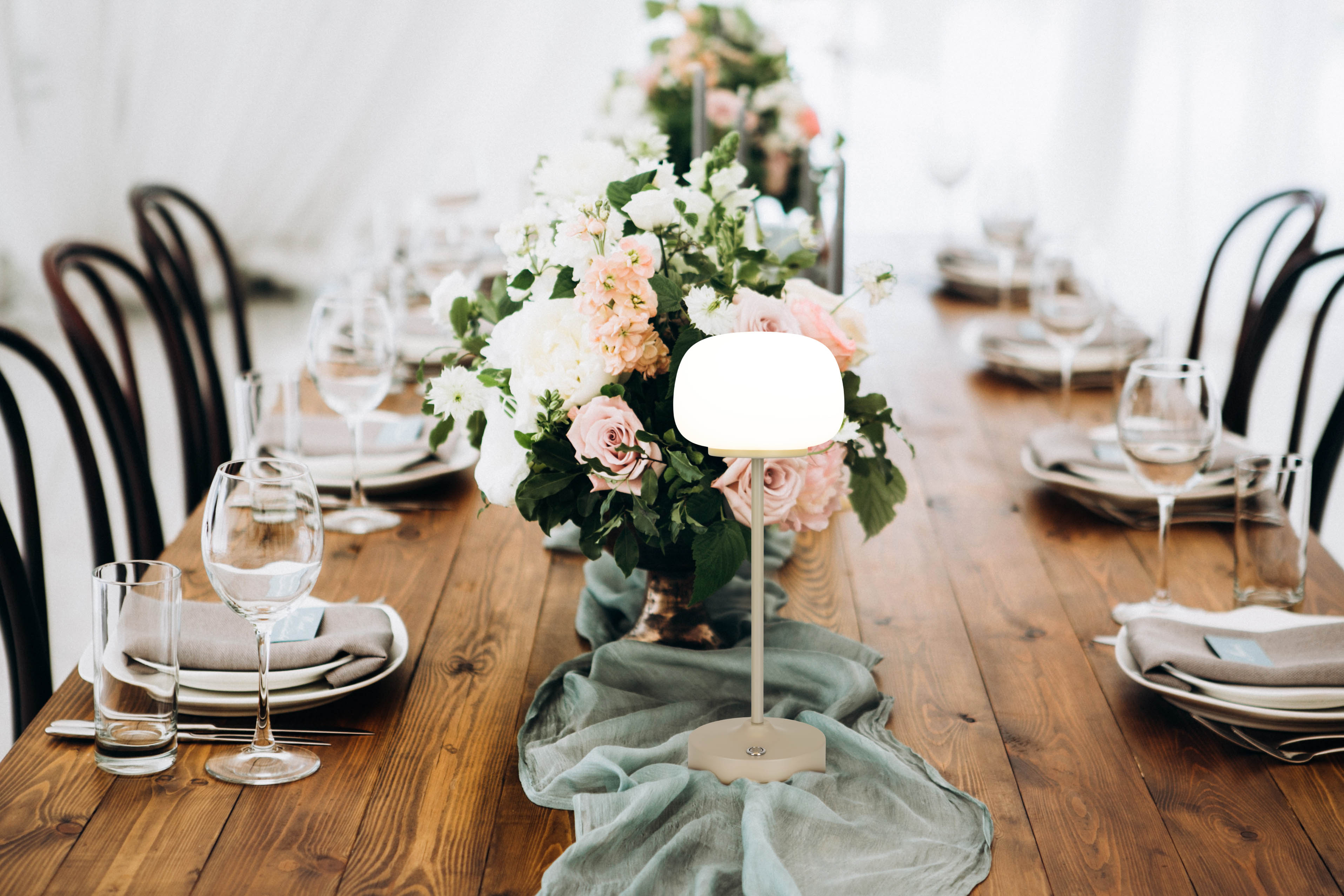Ein Esstisch für einen festlichen Anlass, mit Blumenschmuck und Lampe in der Mitte und Stühlen drumherum.