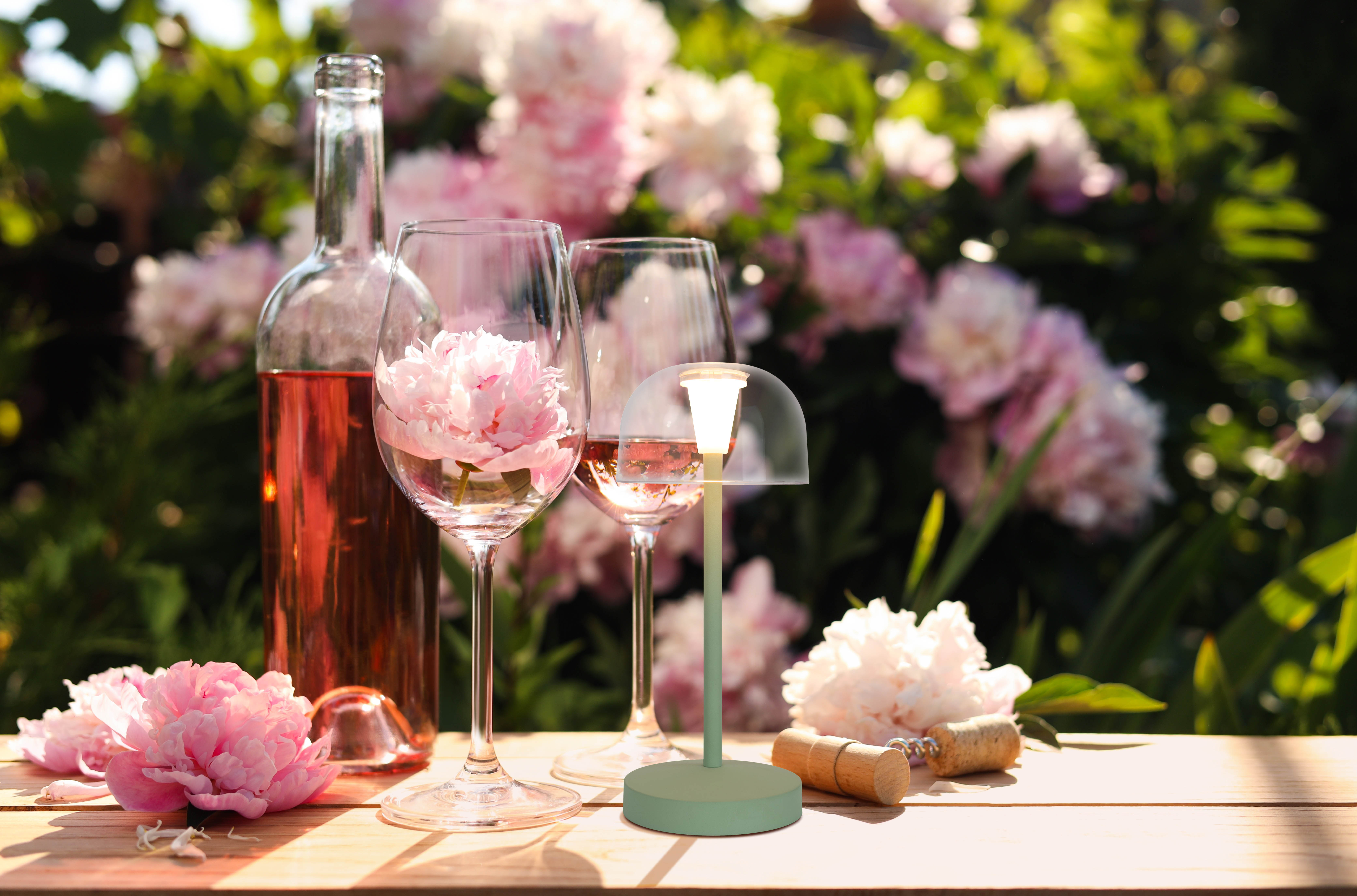 Ein Tisch mit Wein, Gläsern mit Blumen und einer Lampe in einer Gartenszene.