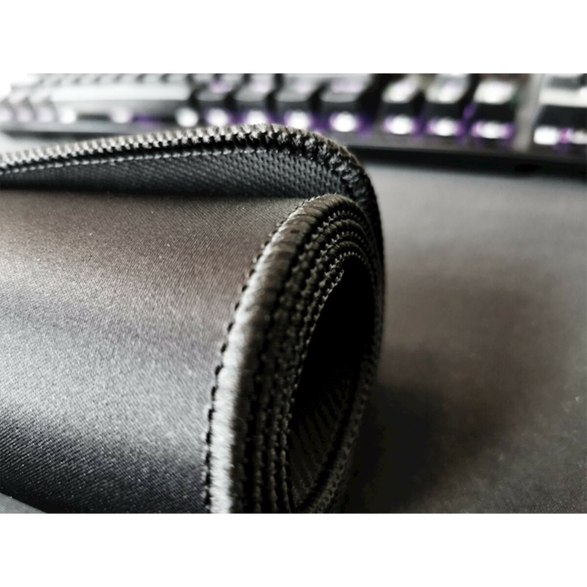 Nahaufnahme eines aufgerollten schwarzen Mauspads mit genähten Kanten. Eine Tastatur ist im Hintergrund unscharf.