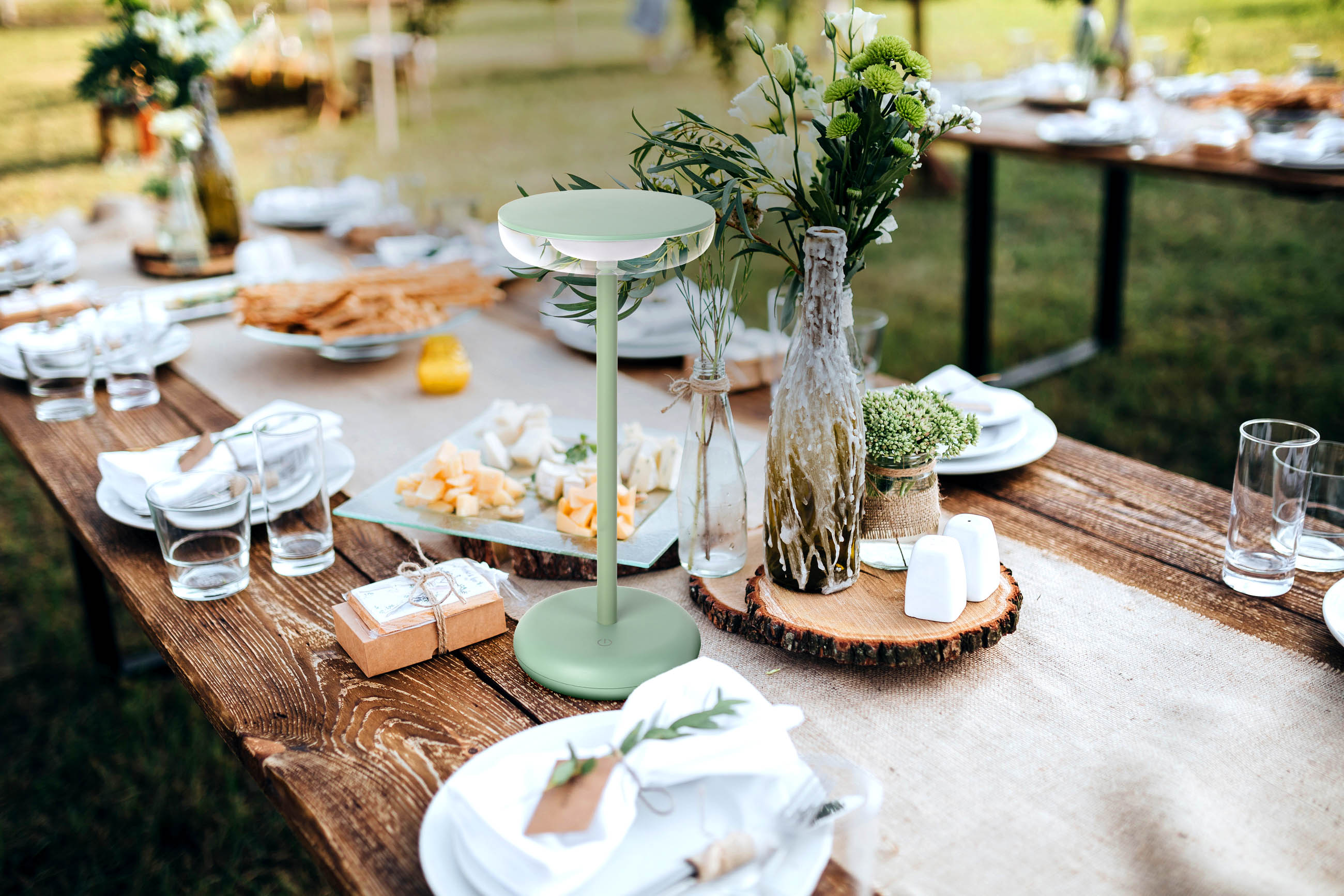 Tisch für Essen im Freien, mit moderner grüner Lampe, Blumen, Käse und Tellern auf einem Holztisch.
