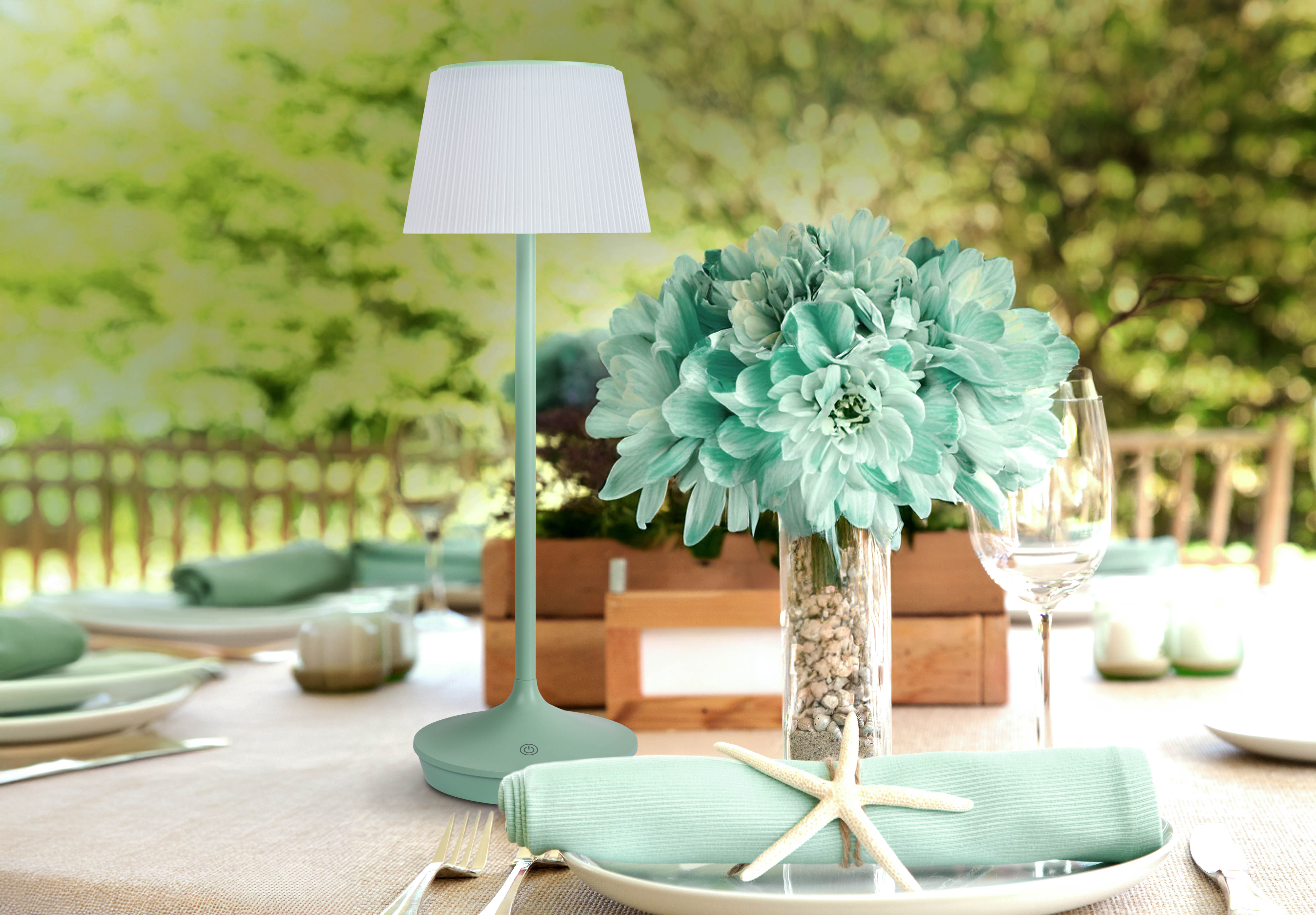 Tischdeko: Lampe, Blumen, Teller und Besteck. Grüner Hintergrund mit Bäumen.