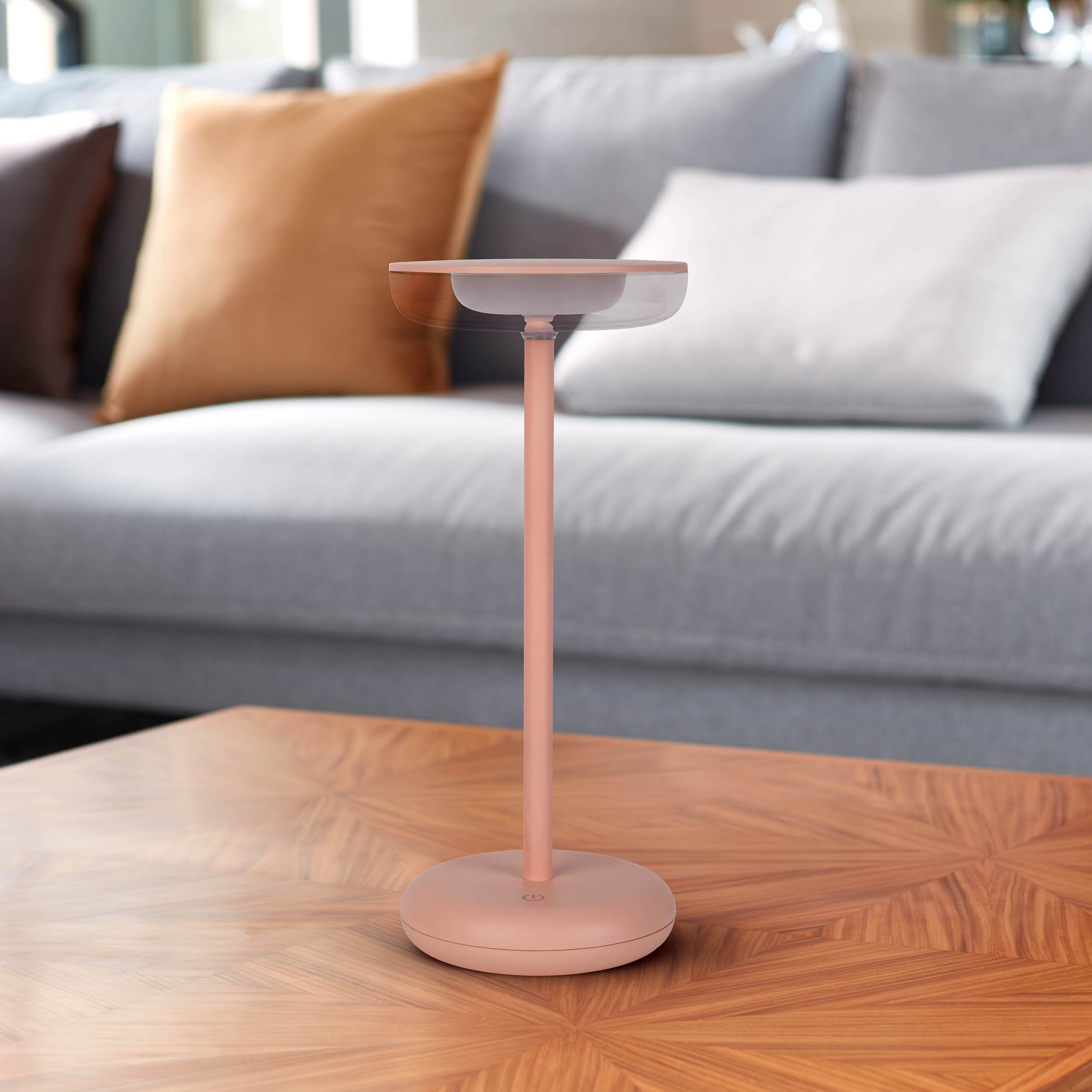 Eine rosa Lampe auf einem Holztisch, mit einem grauen Sofa im Hintergrund.