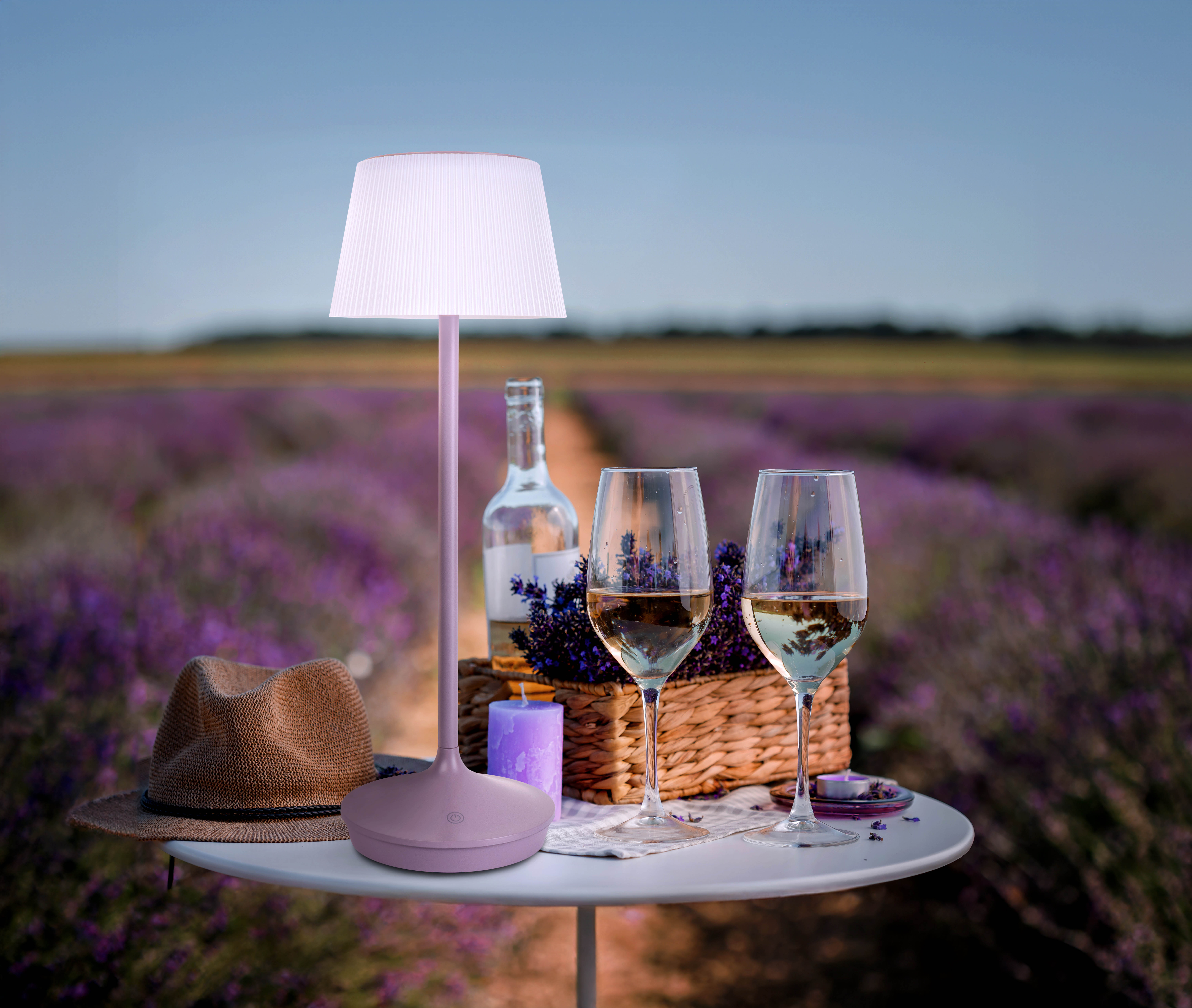Ein Tisch mit zwei Weingläsern, einer Lampe, einer Flasche, einem Hut und einem Lavendelfeld im Hintergrund.