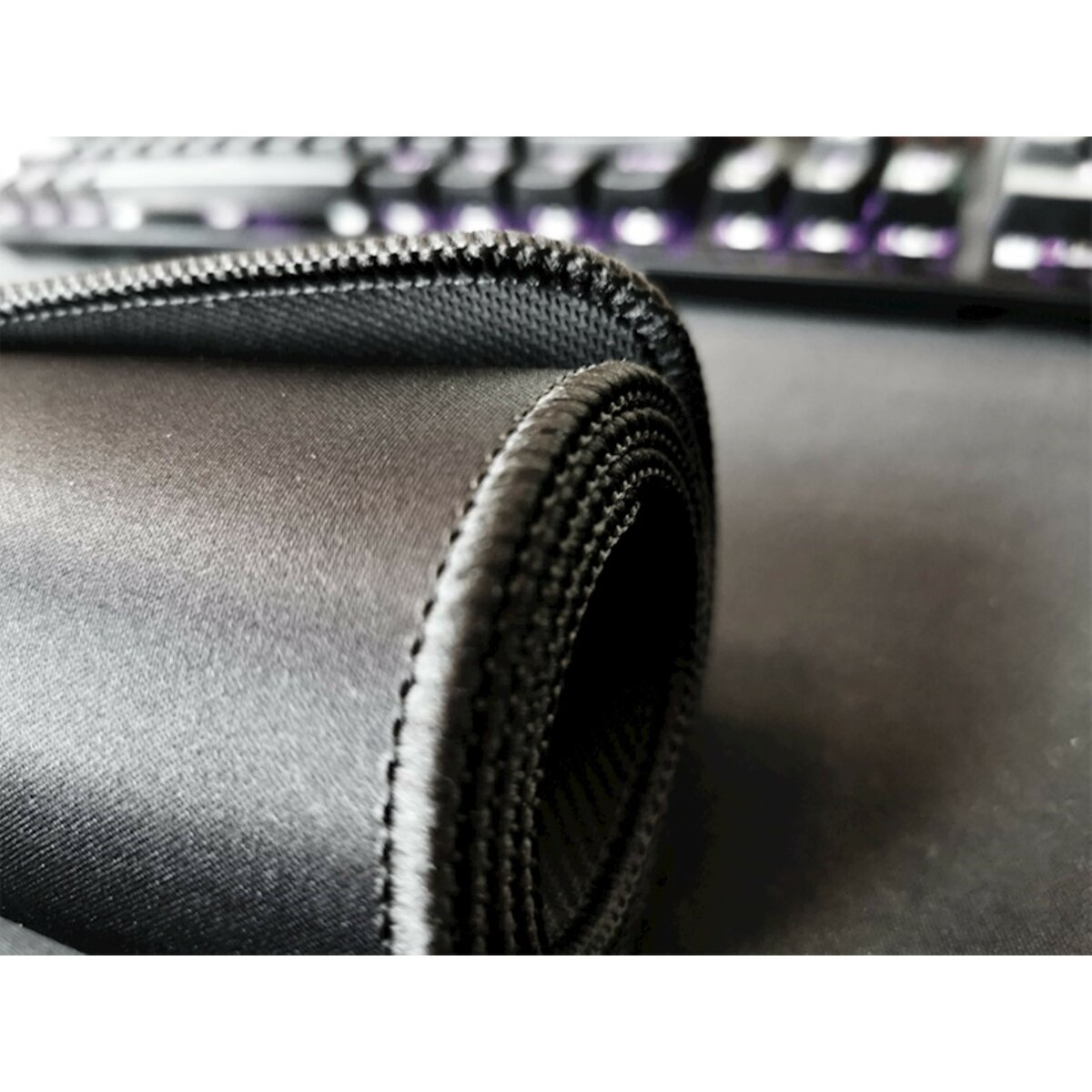 Ein schwarzes Mousepad mit gerolltem Rand und eine Tastatur im Hintergrund.