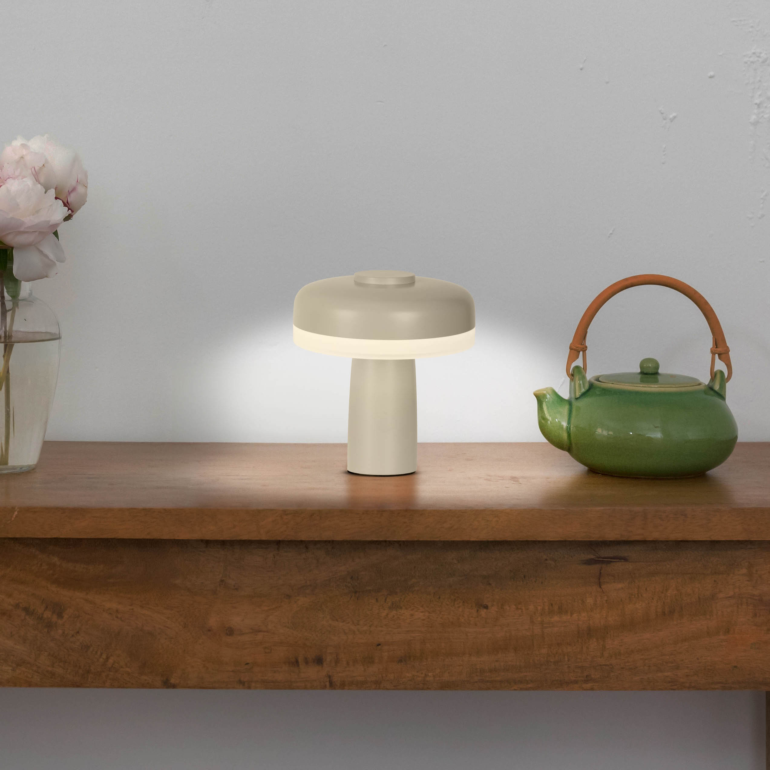 Eine beige pilzförmige Lampe auf einem Holztisch, eine Vase mit Blumen und eine grüne Teekanne.