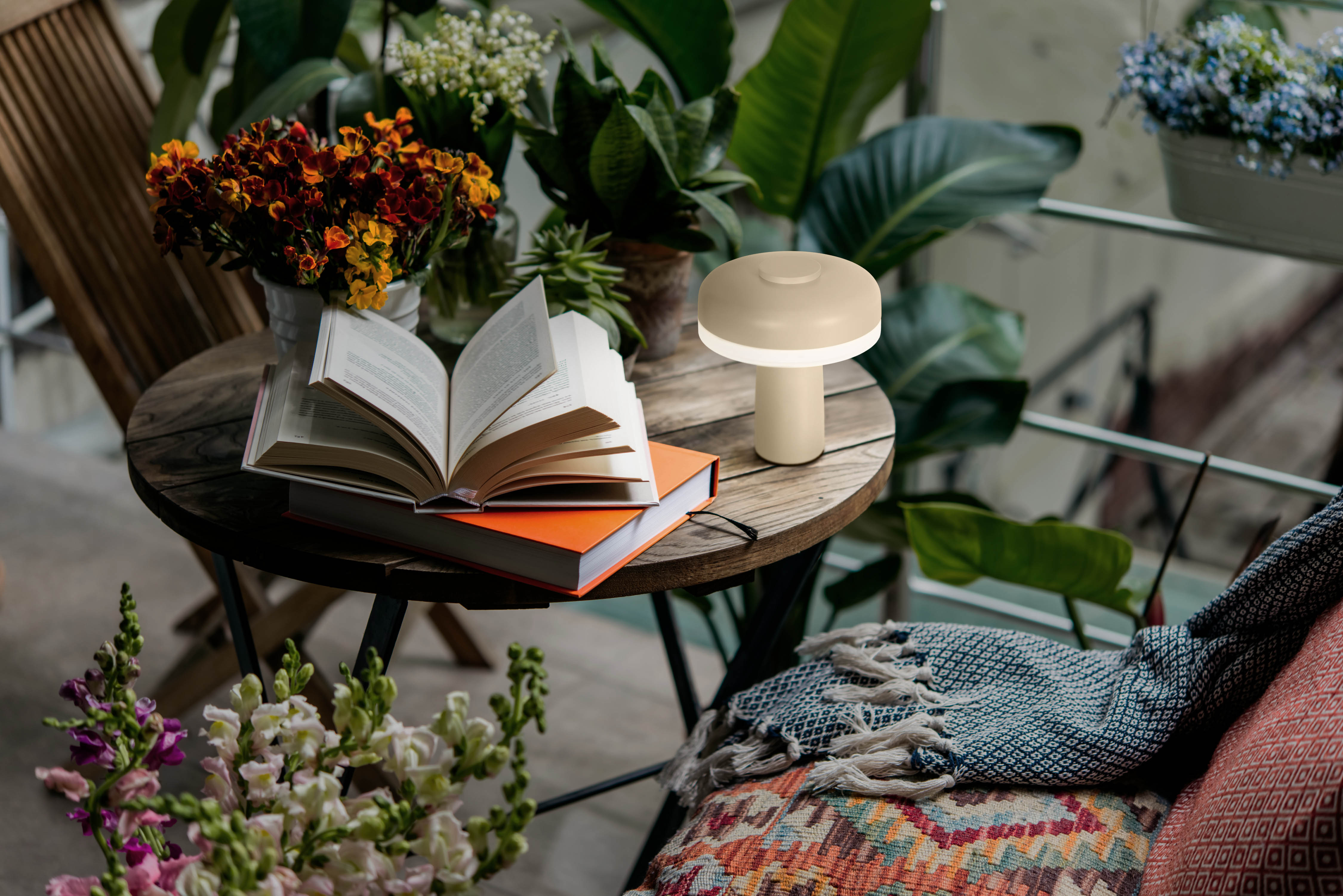 Tisch mit Büchern, Lampe und Blumen. Hintergrund mit Pflanzen und bunter Decke.