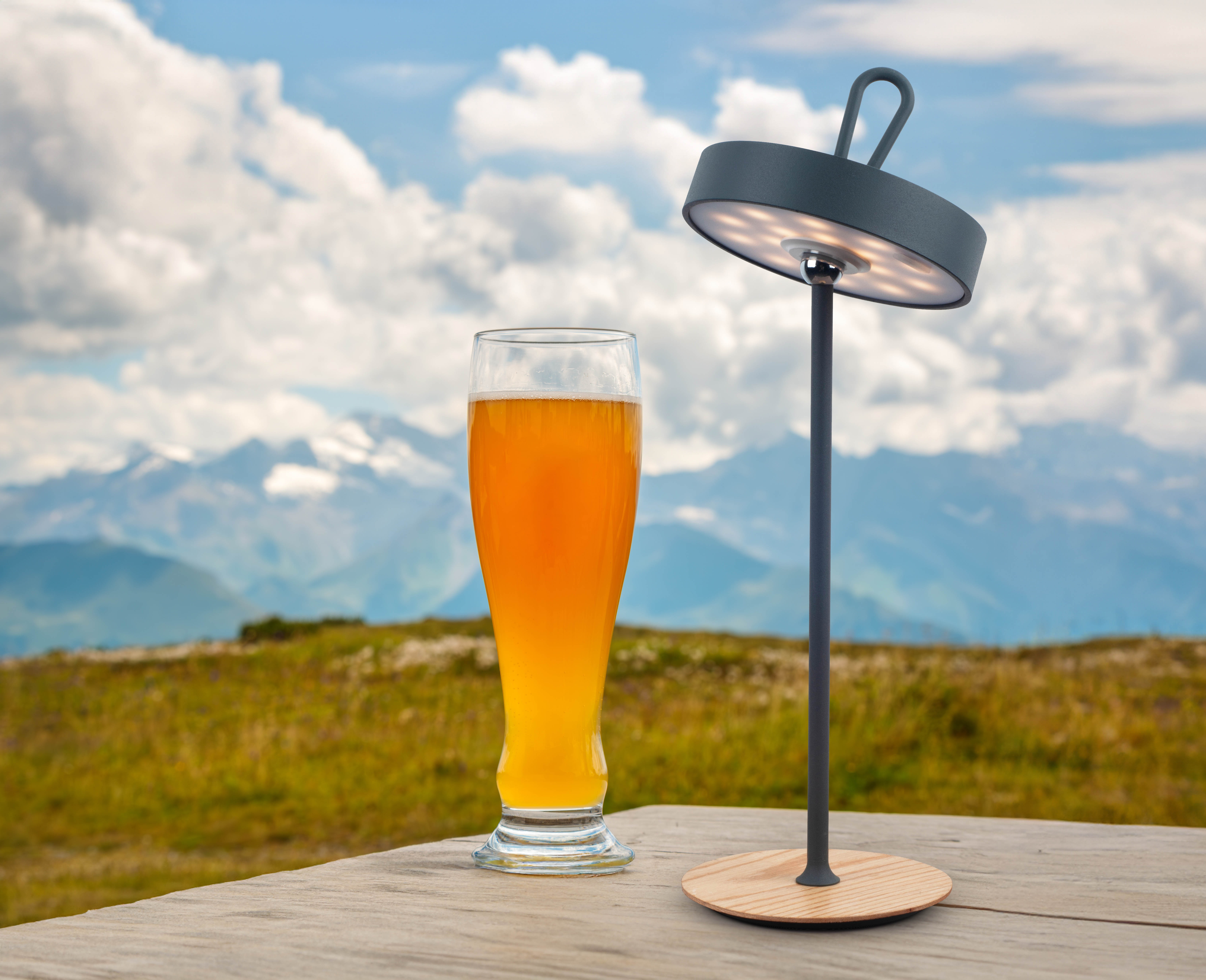 Szene im Freien mit Bierglas, Lampe und Bergen im Hintergrund. Holztisch.