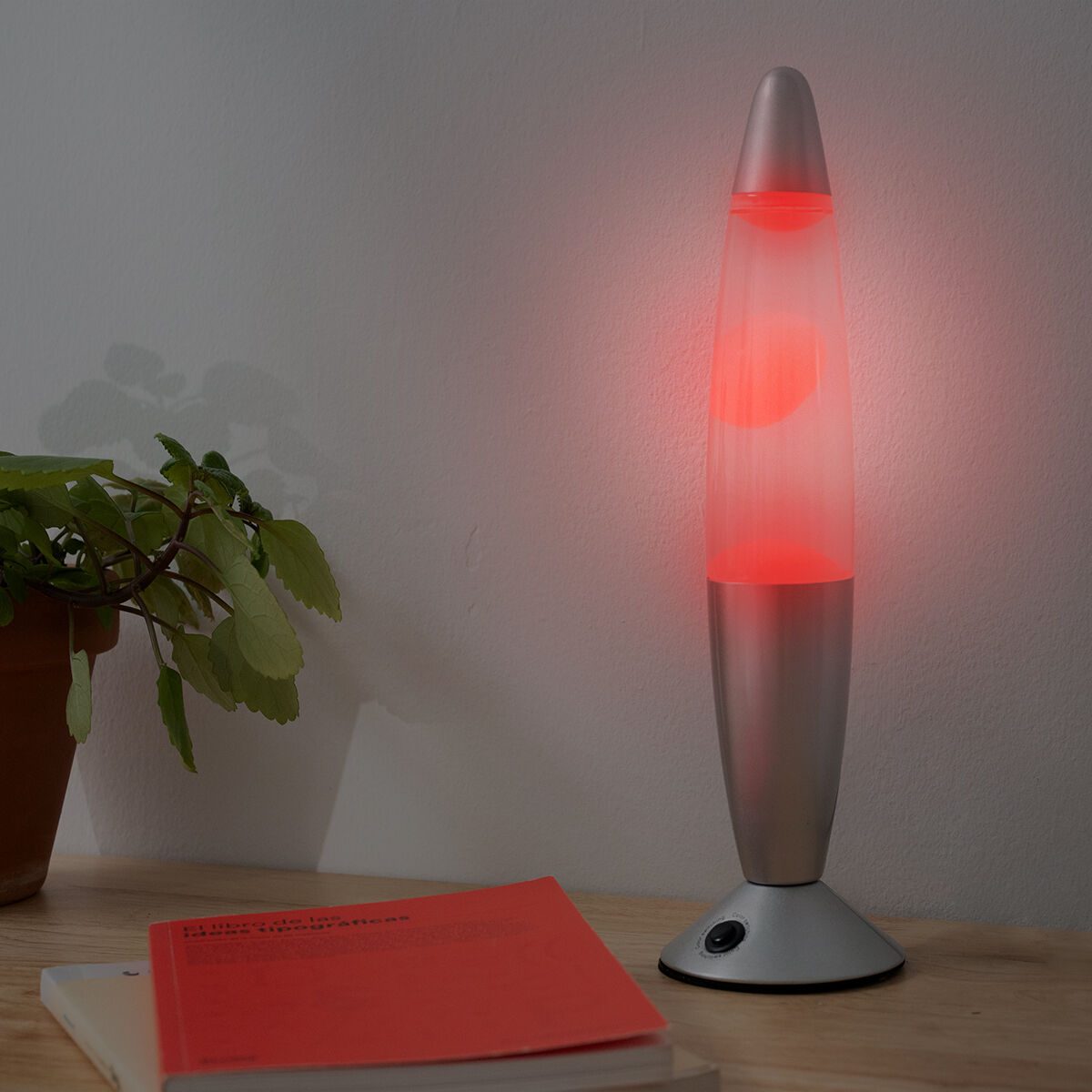 Lámpara de lava con base plateada y tubo transparente con lava roja. Está sobre una mesa de madera.