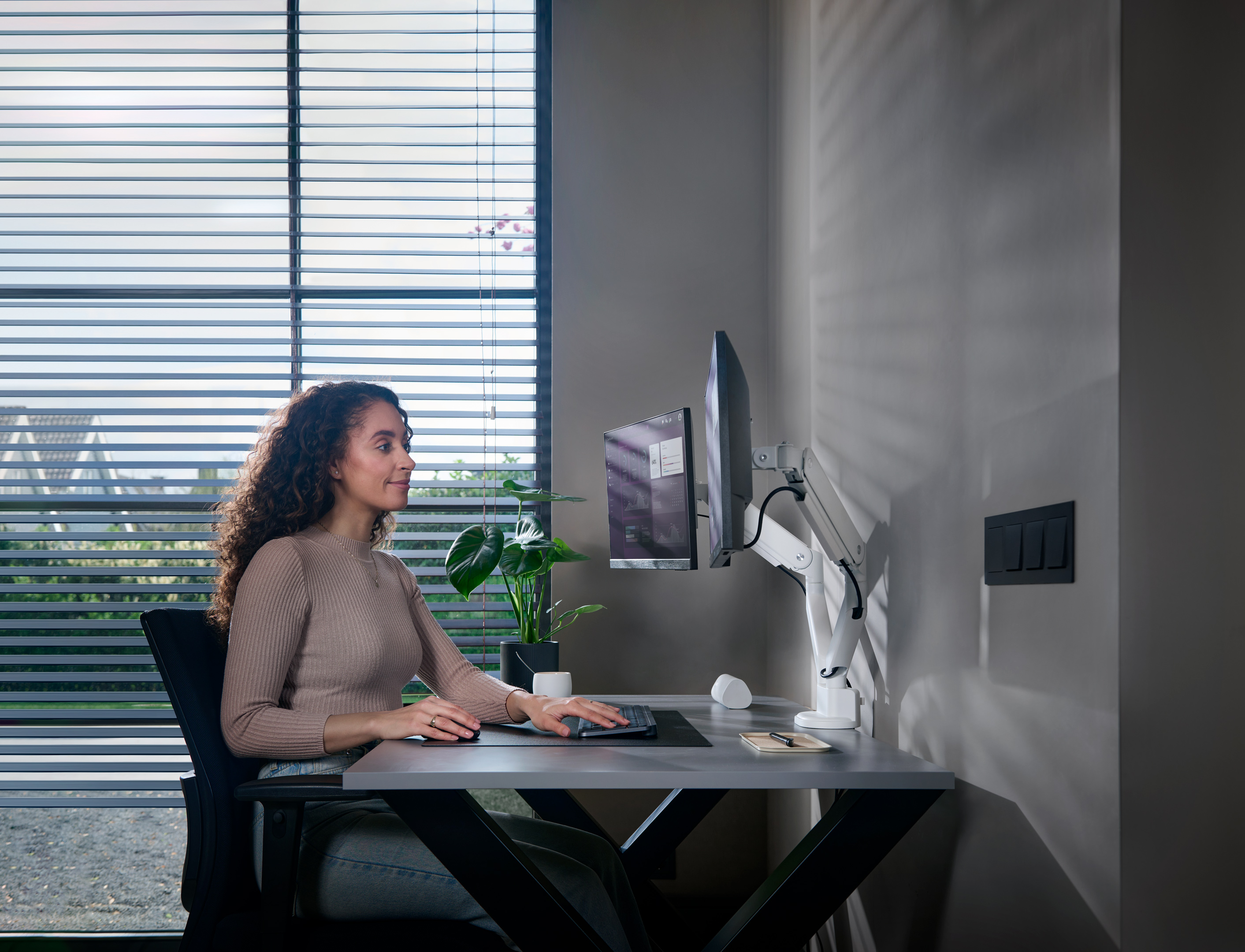 Kobieta siedzi przy biurku z dwoma monitorami, pracując. Światło słoneczne prześwituje przez żaluzje w tle.