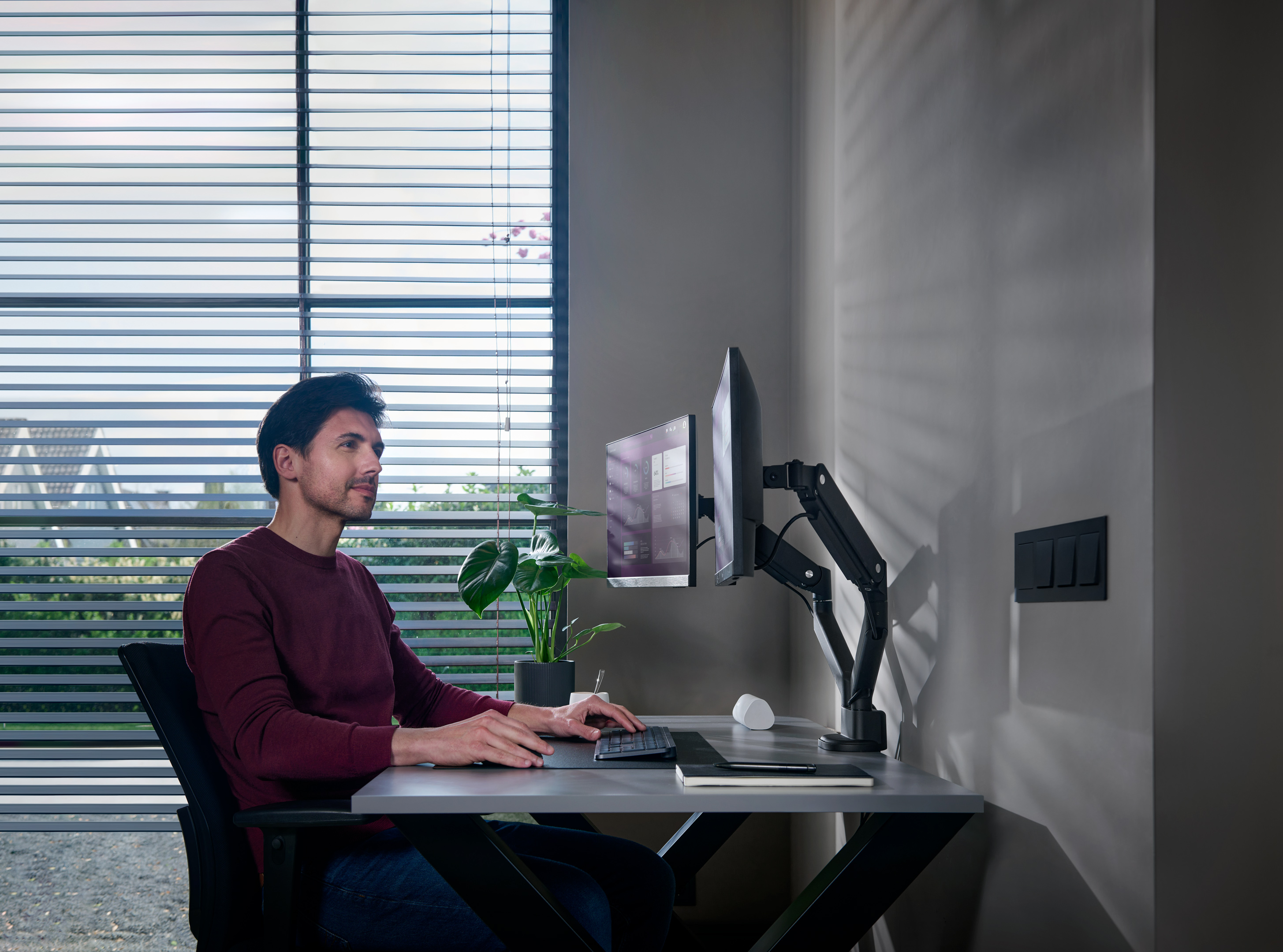 Man working at desk with dual monitors.  Window blinds, plant, and wall with light switch.