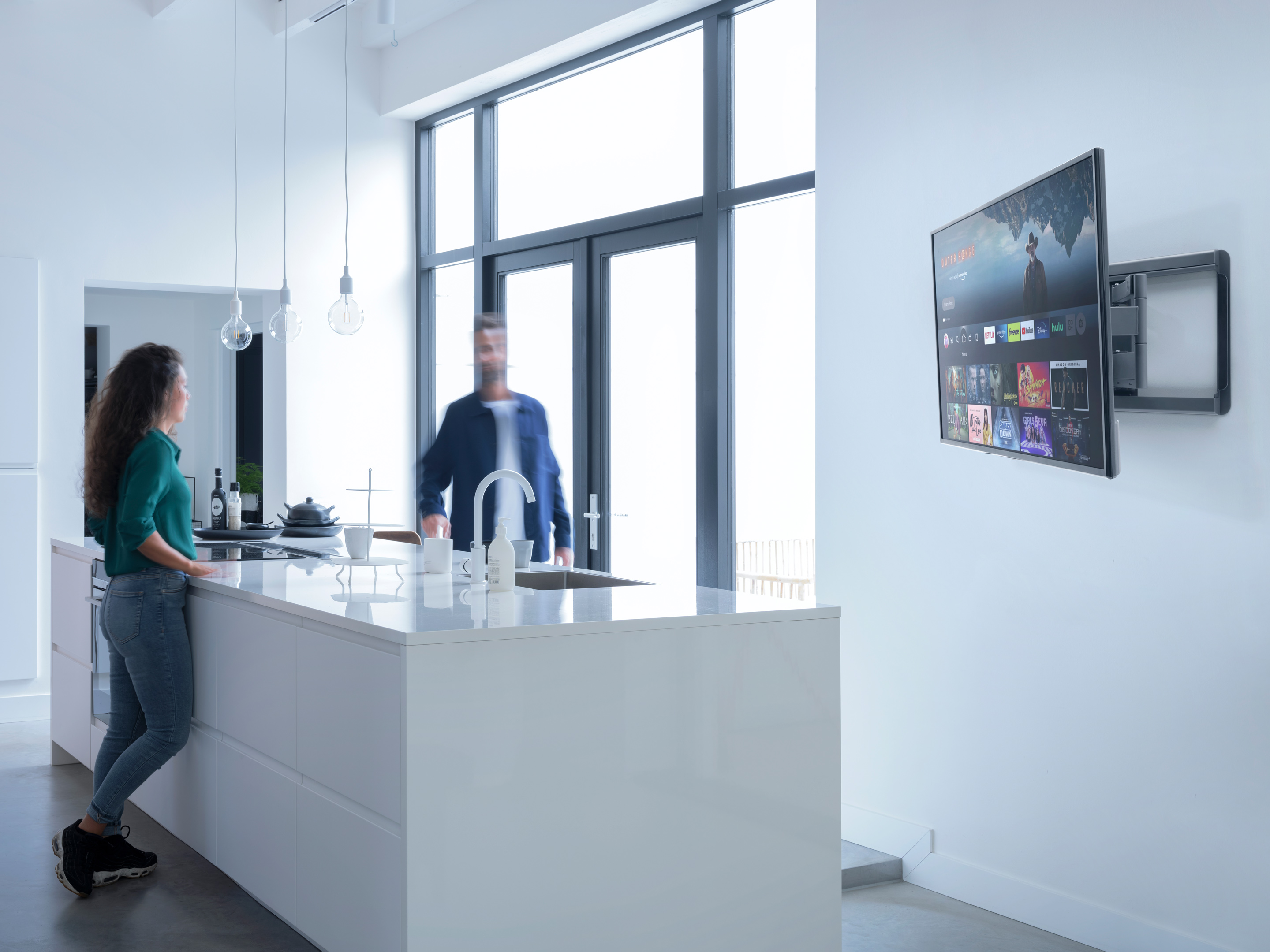 A person stands in a kitchen looking at a TV mounted on the wall. A man walks in the background.
