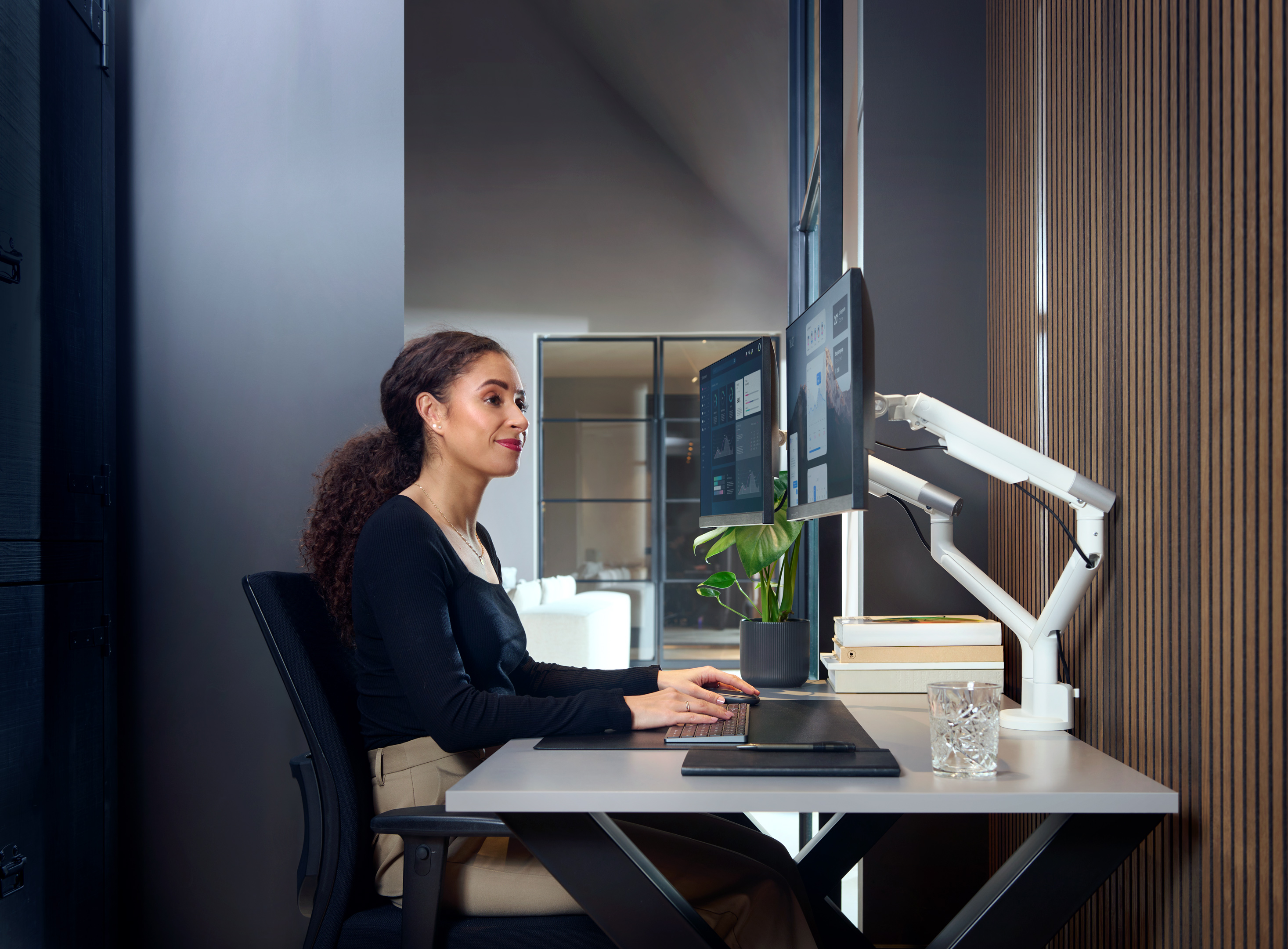Vrouw werkt aan een bureau met dubbele monitoren, toetsenbord en plant. Moderne kantooromgeving met houten panelen.
