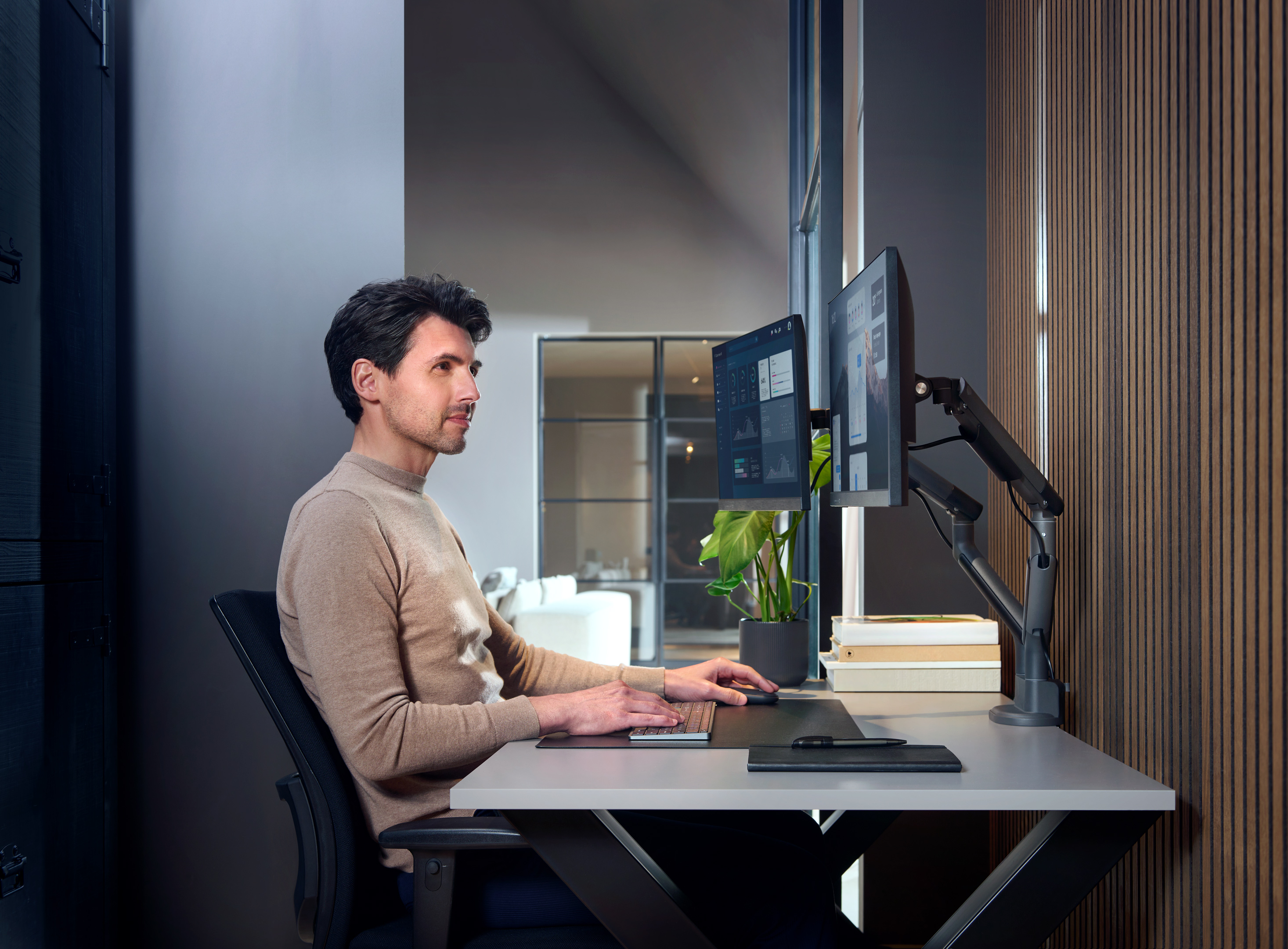 Mężczyzna pracuje przy biurku z dwoma monitorami i ramieniem do monitora. Widoczna roślina.