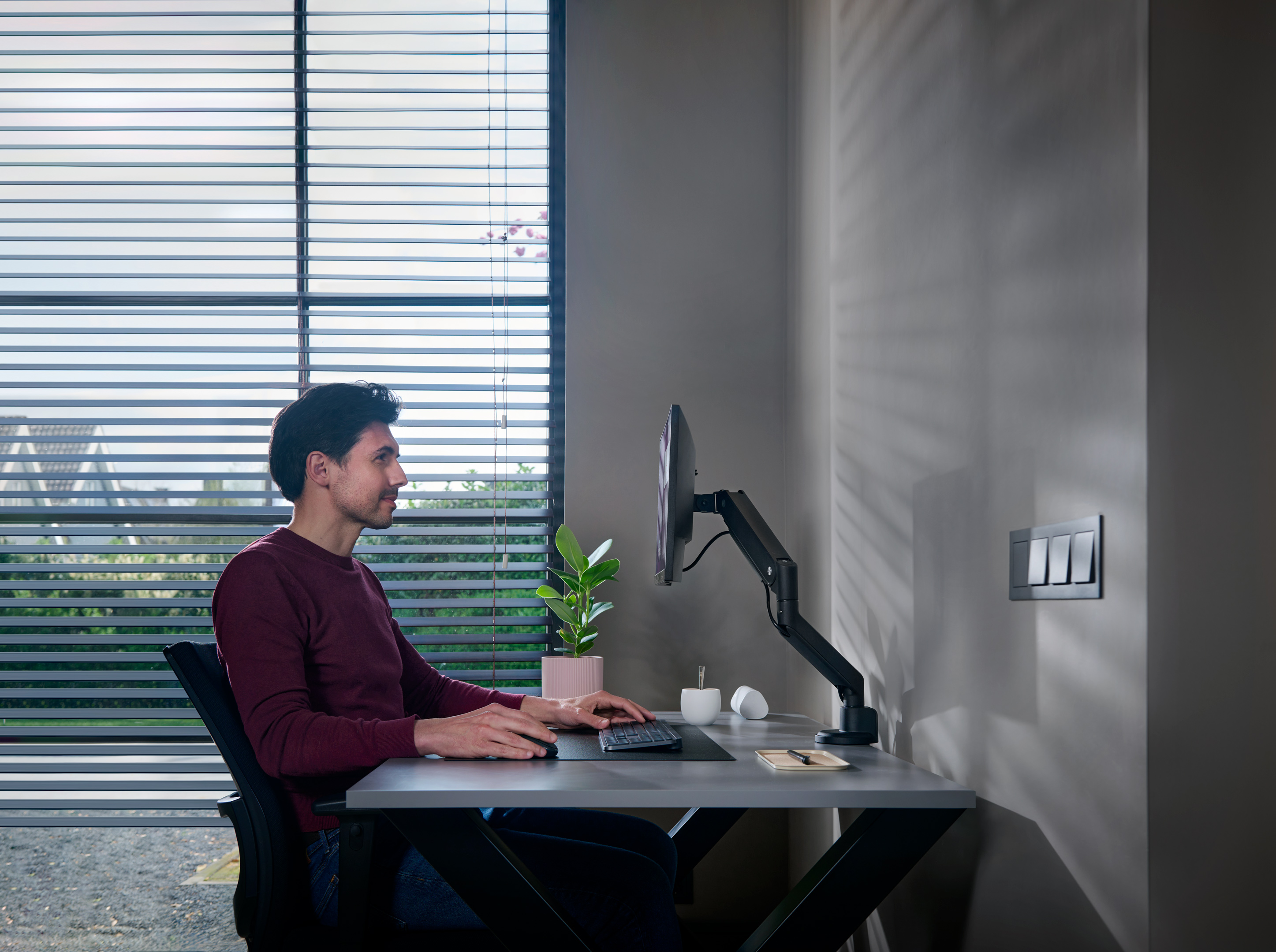 Mężczyzna pracuje przy biurku z monitorem, rośliną, klawiaturą i szarą ścianą z włącznikami.