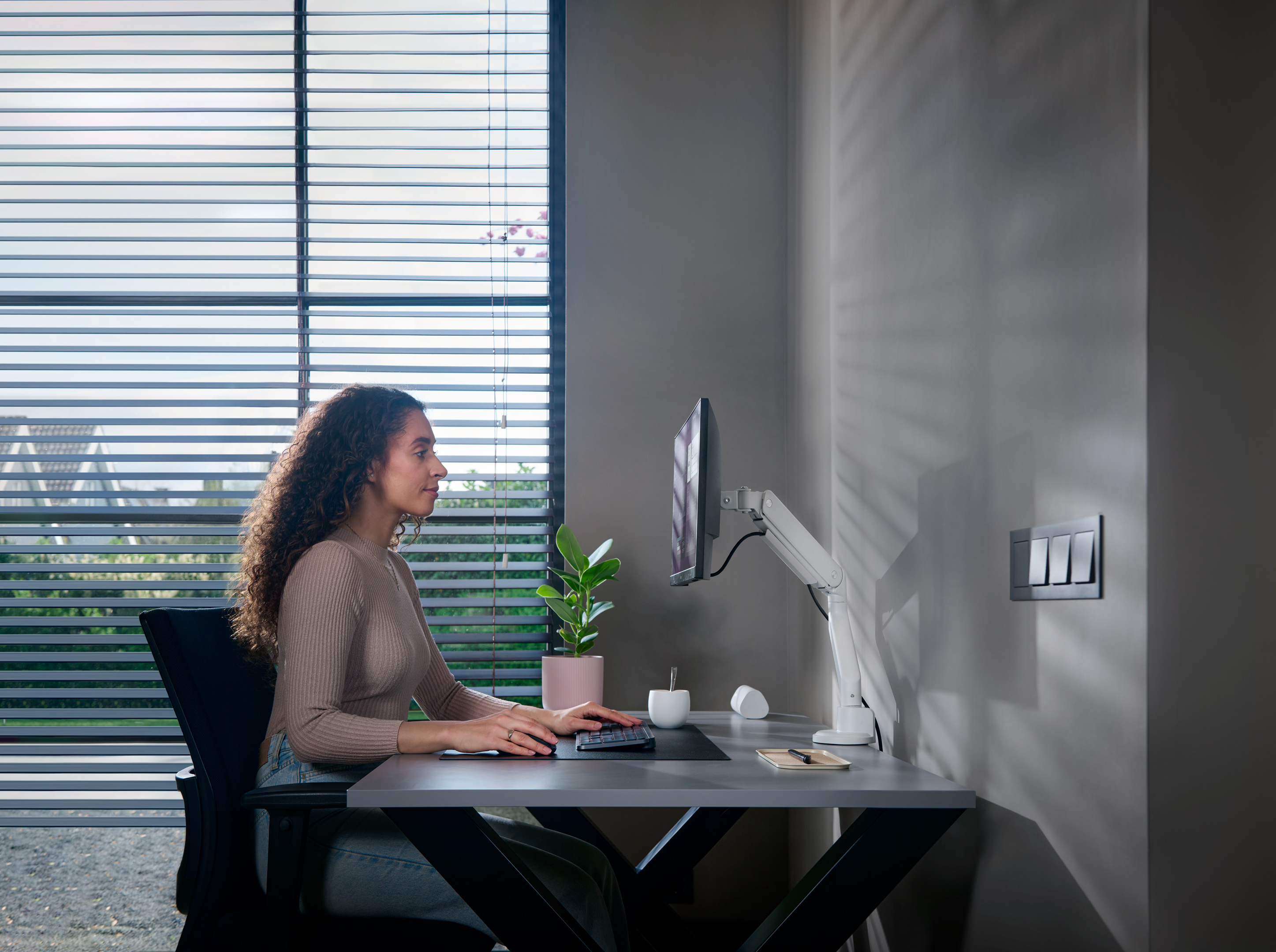 Frau am Schreibtisch mit Computer, Monitor an weißem Arm, Pflanze, Fenster.