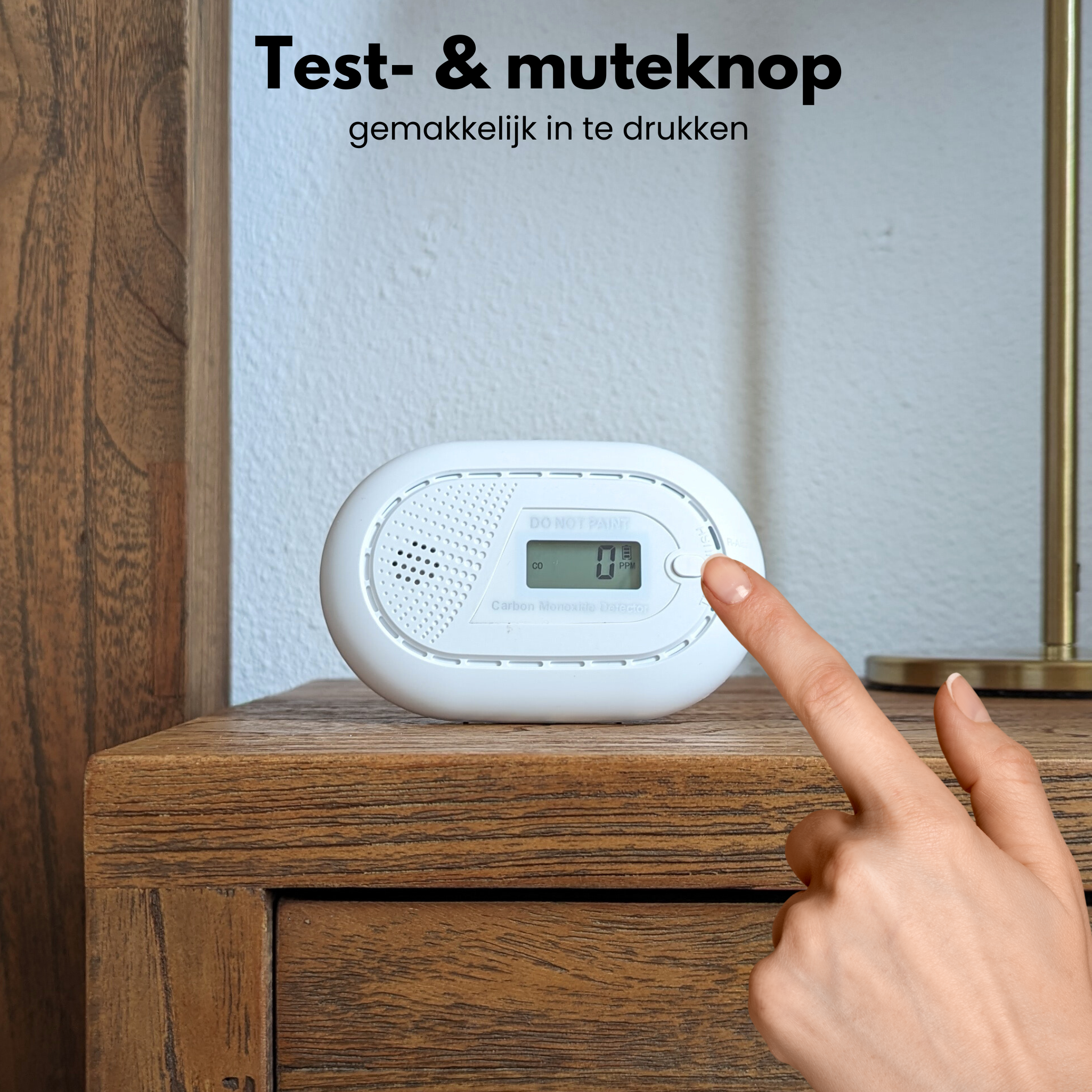 A white carbon monoxide detector on a wooden table. A finger presses a button on the detector. Text: Test & mute button.