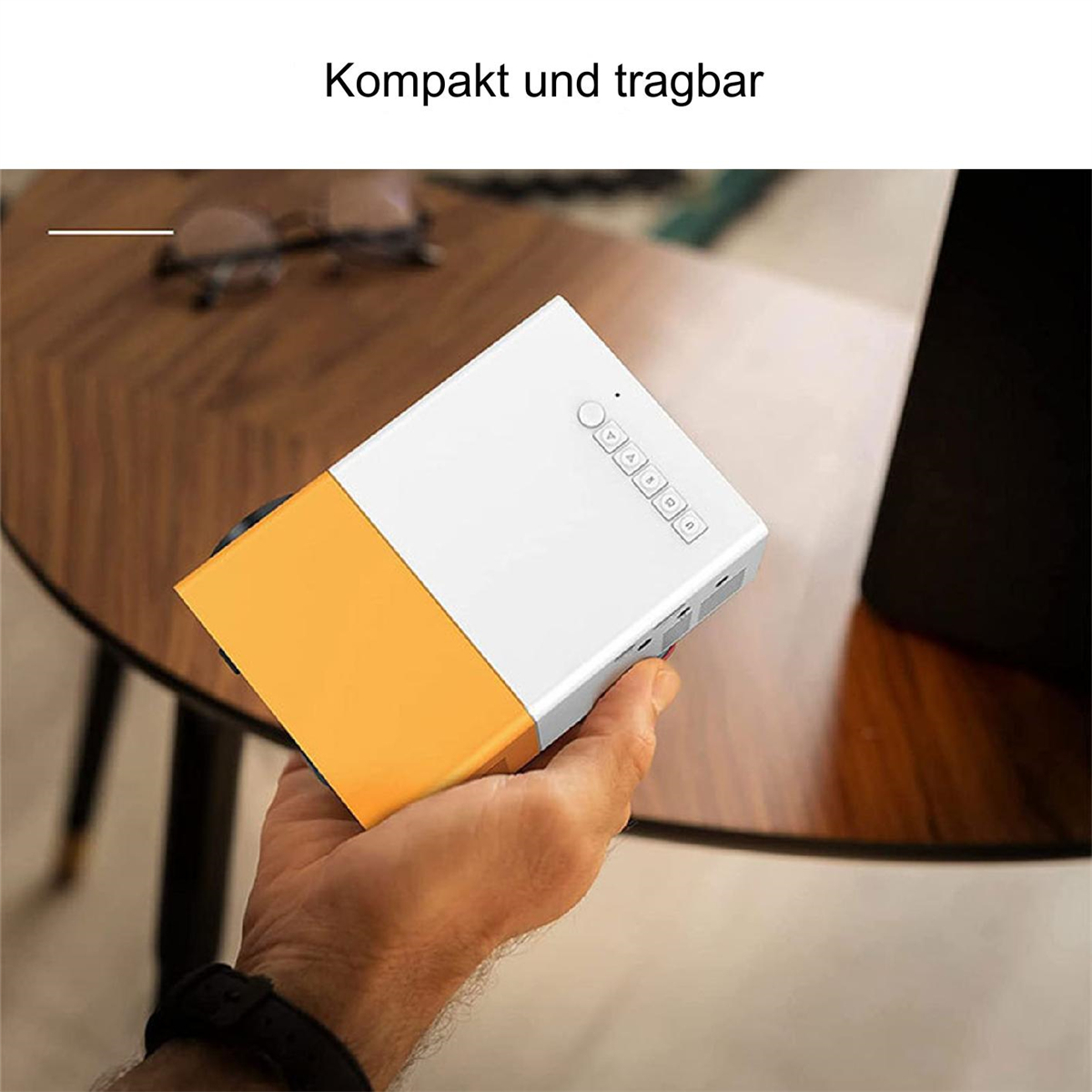 Hand hält weißen und orangenen Mini-Projektor. Knöpfe und Tisch vorhanden.