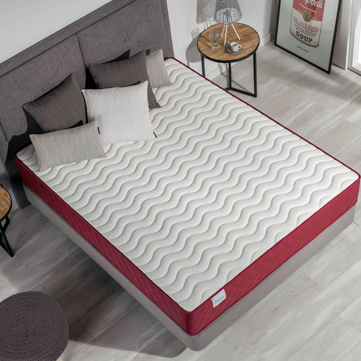 Cama con colchón, cabecero gris y almohadas. Borde rojo en el colchón.