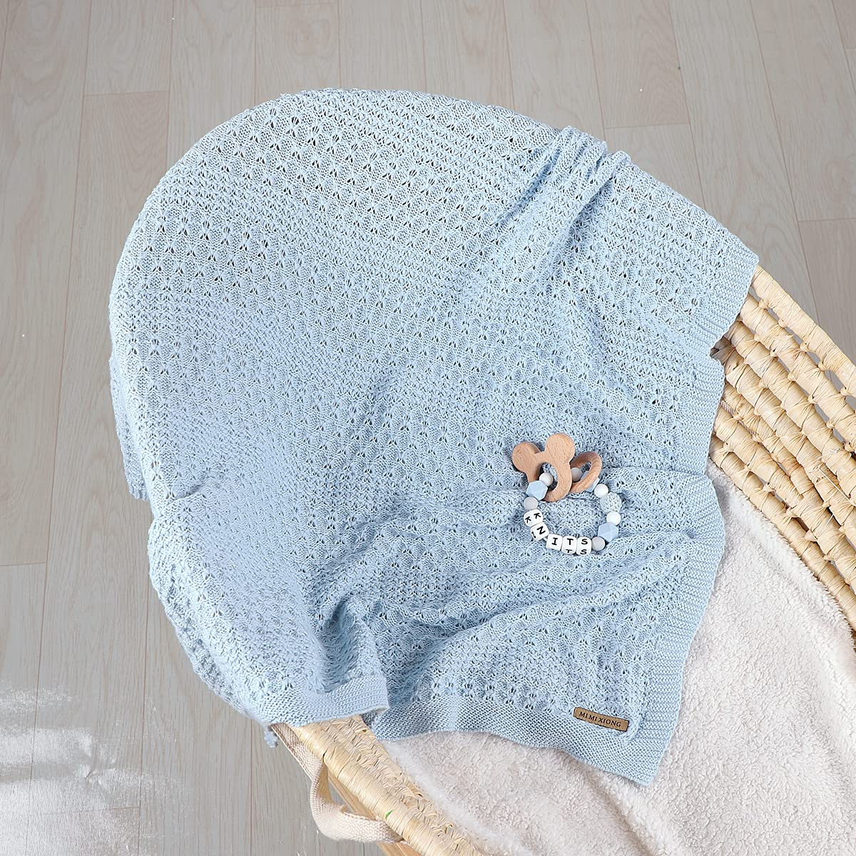 Eine hellblaue gestrickte Babydecke mit Holzspielzeug und Schnuller in einem Weidenkorb.