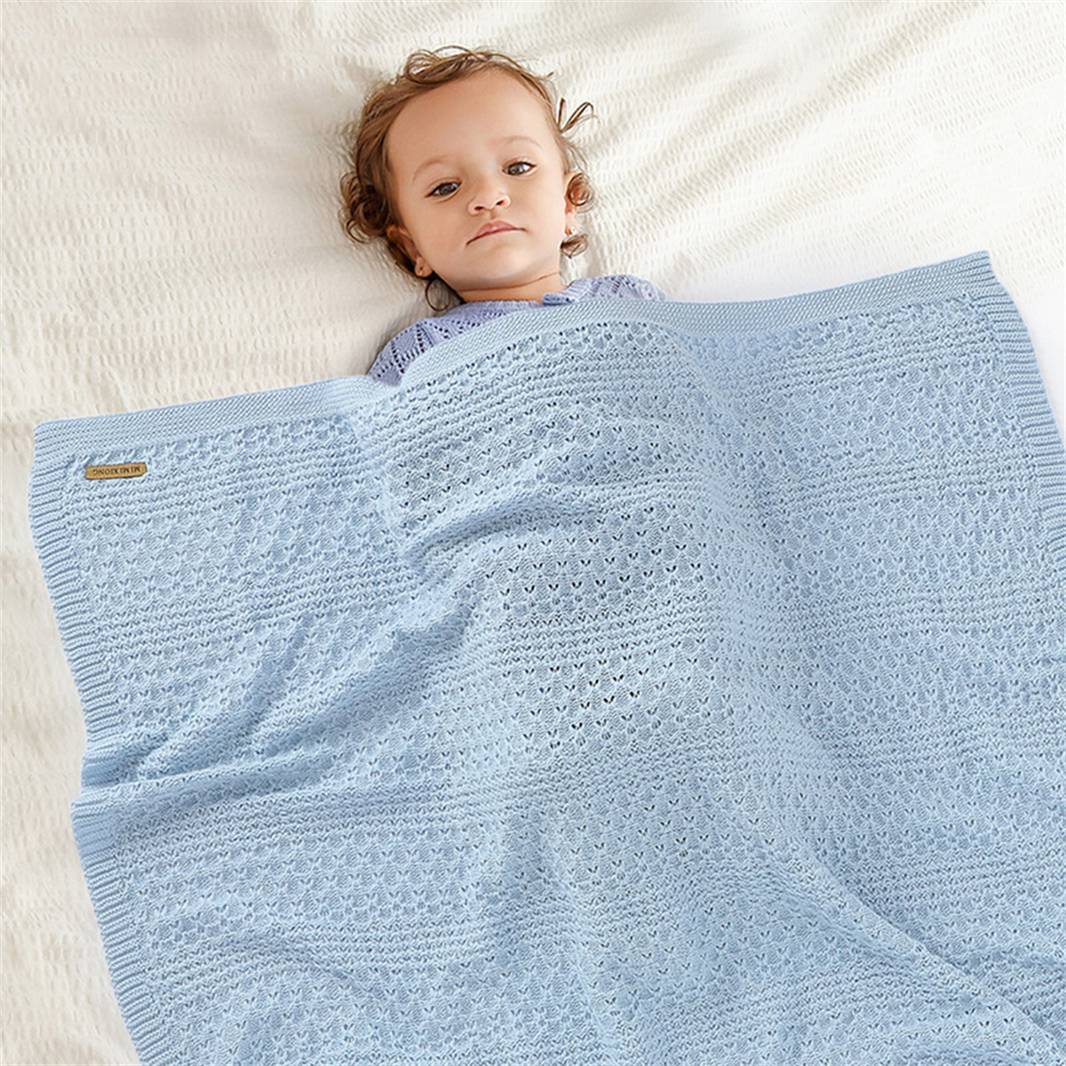 Ein Baby liegt auf einem Bett, bedeckt von einer blauen Strickdecke. Das Baby blickt in die Kamera.