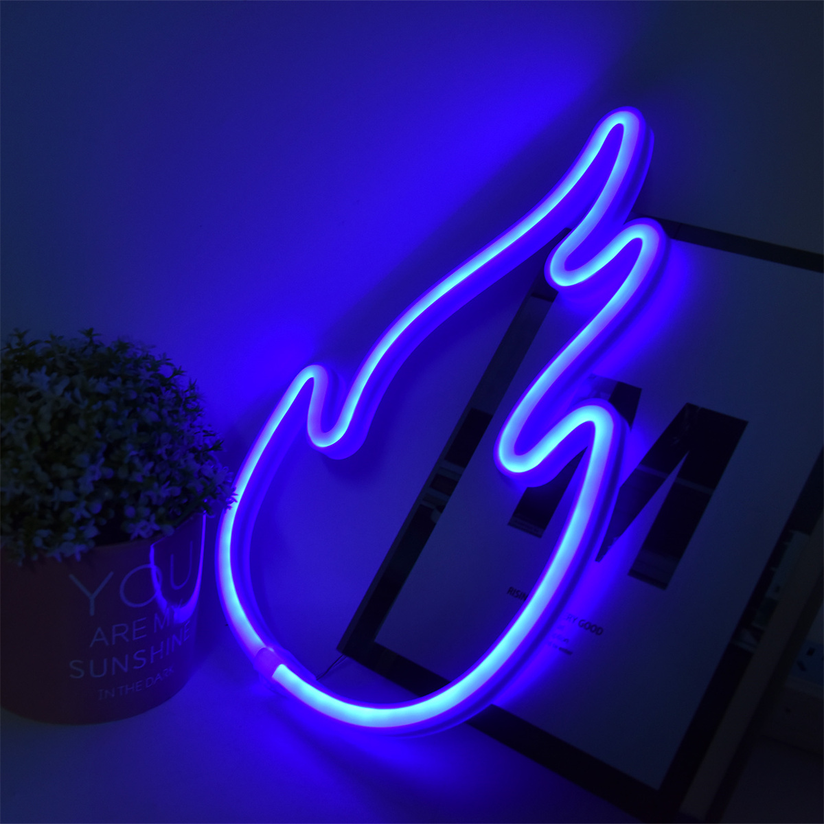 Ein blaues Neonflammenzeichen. Es steht auf einem Tisch mit einer Pflanze und einem Bilderrahmen.