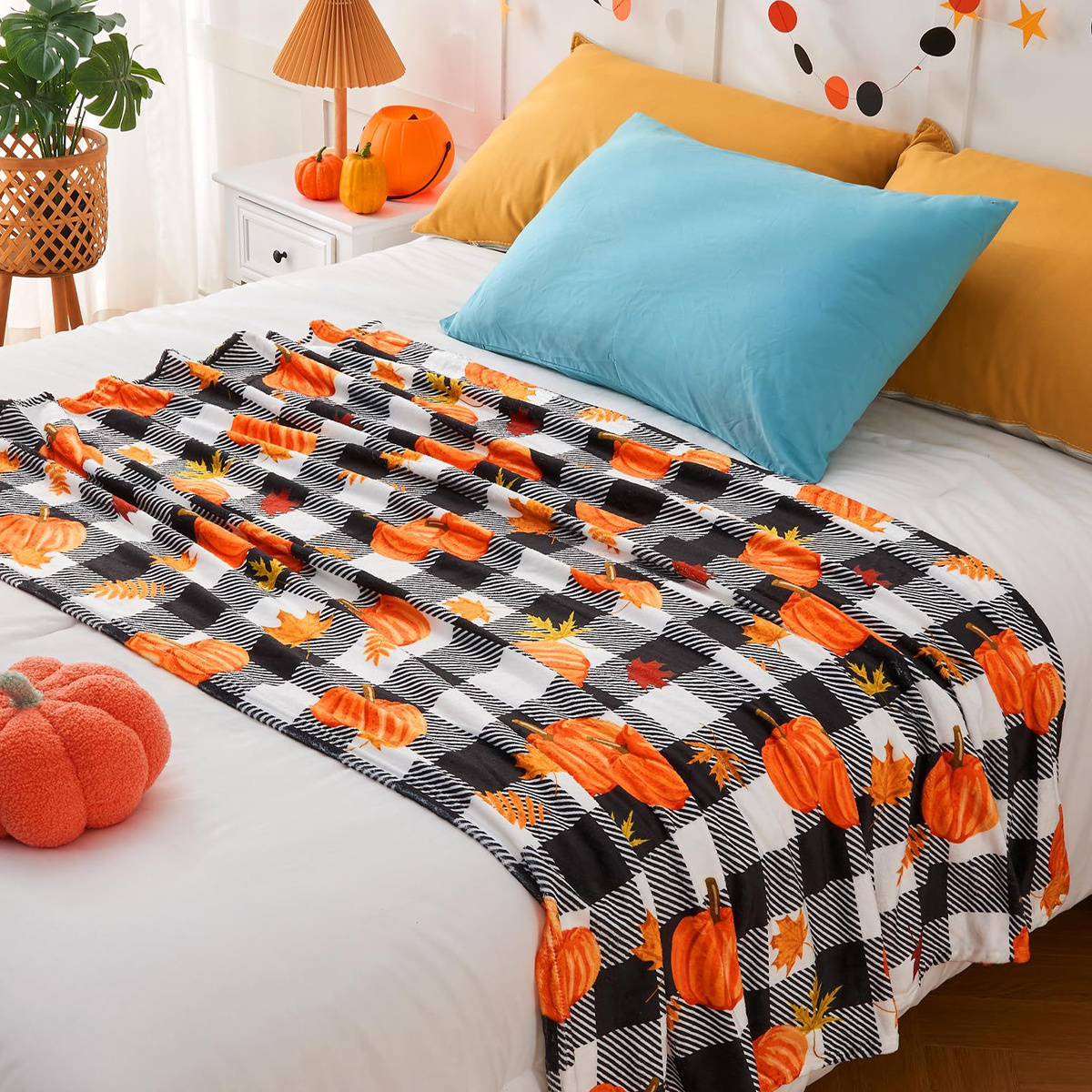 Bett mit Decke mit Kürbissen und Blättern. Es gibt orangefarbene Kissen, ein blaues Kissen und einen Kürbis.