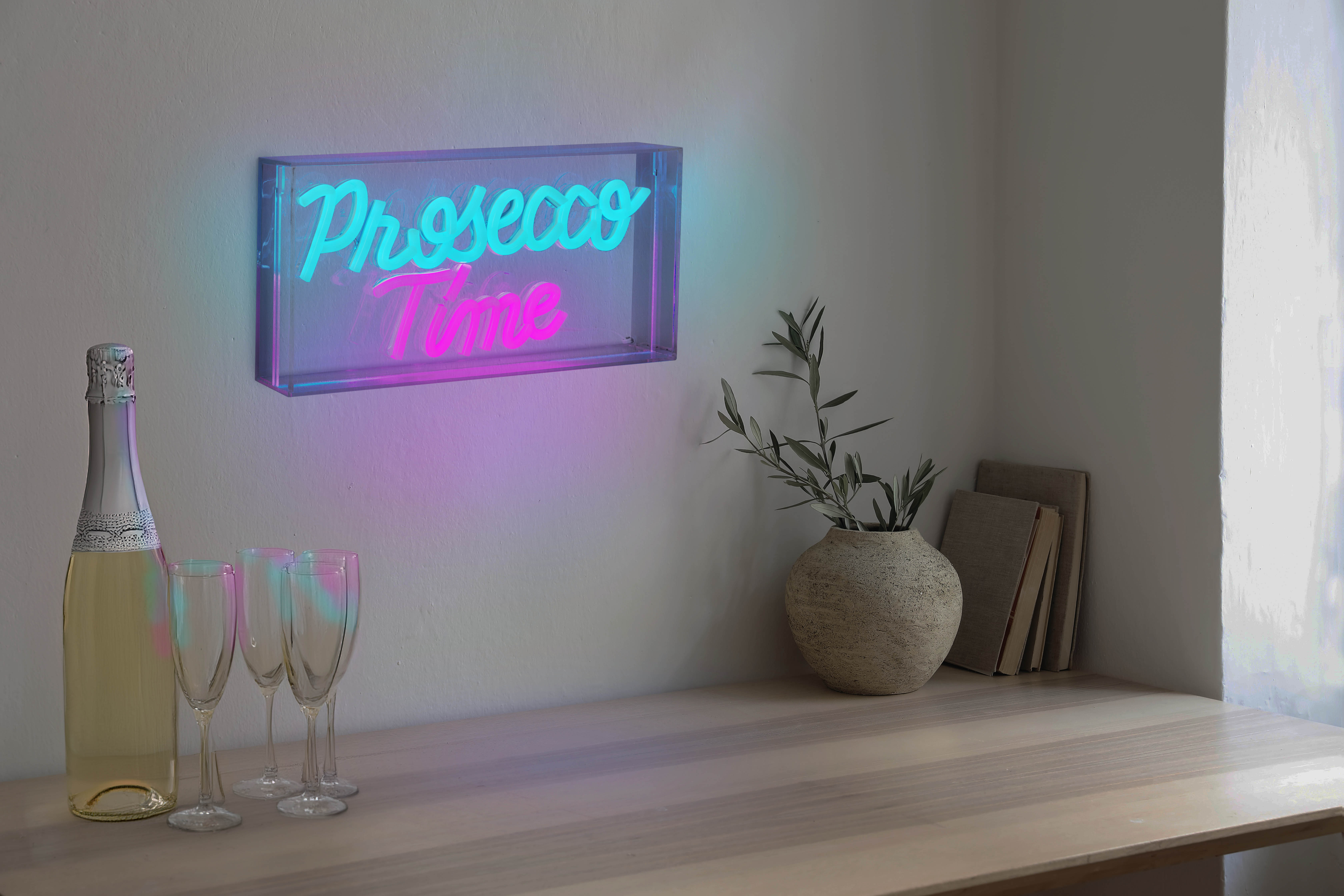 Ein Neonschild mit der Aufschrift 'Prosecco Time' über einem Tisch mit Gläsern, Flasche und Vase.