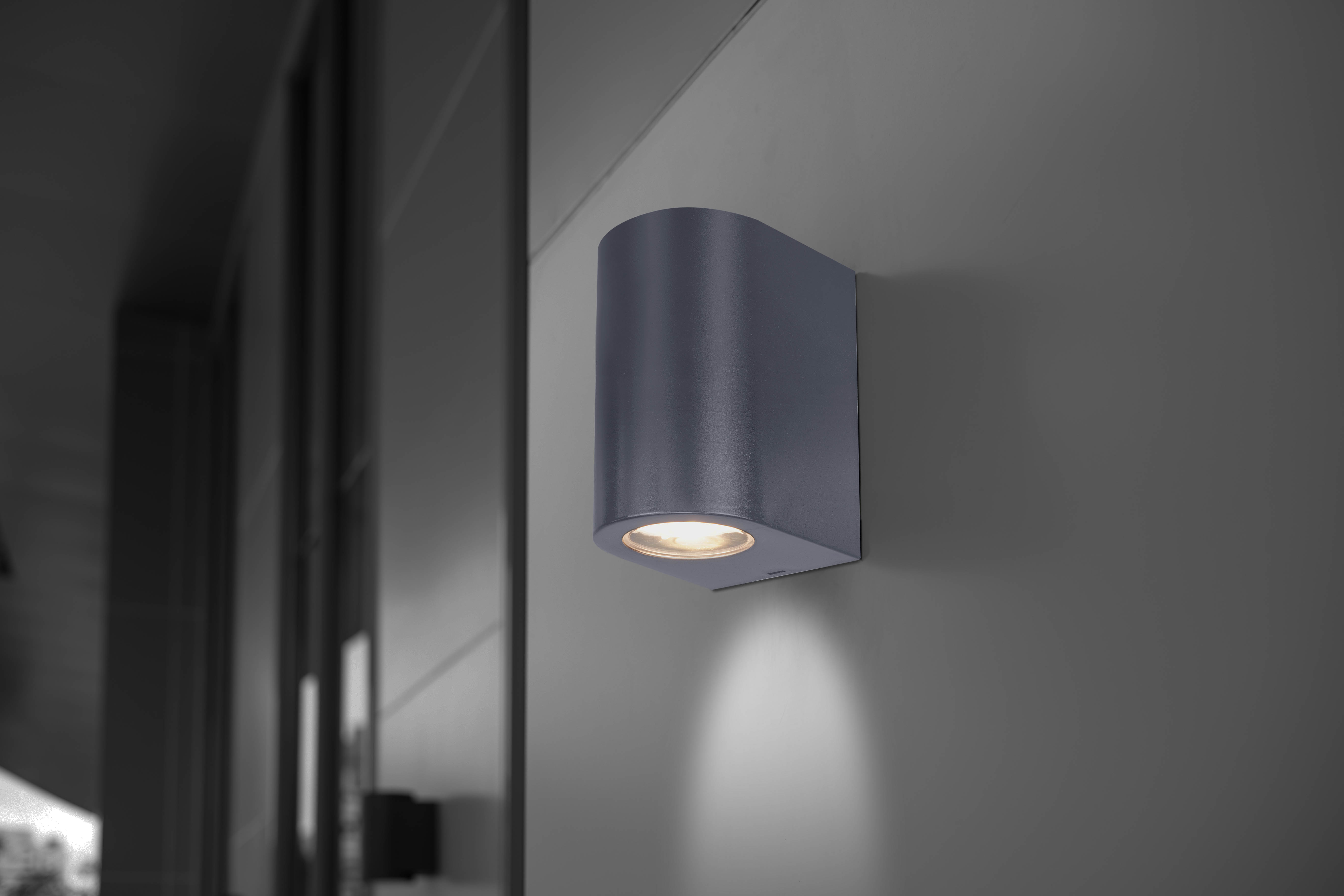 Eine grau an der Wand montierte Außenleuchte. Das Licht ist dunkelgrau, an der Wand befestigt.