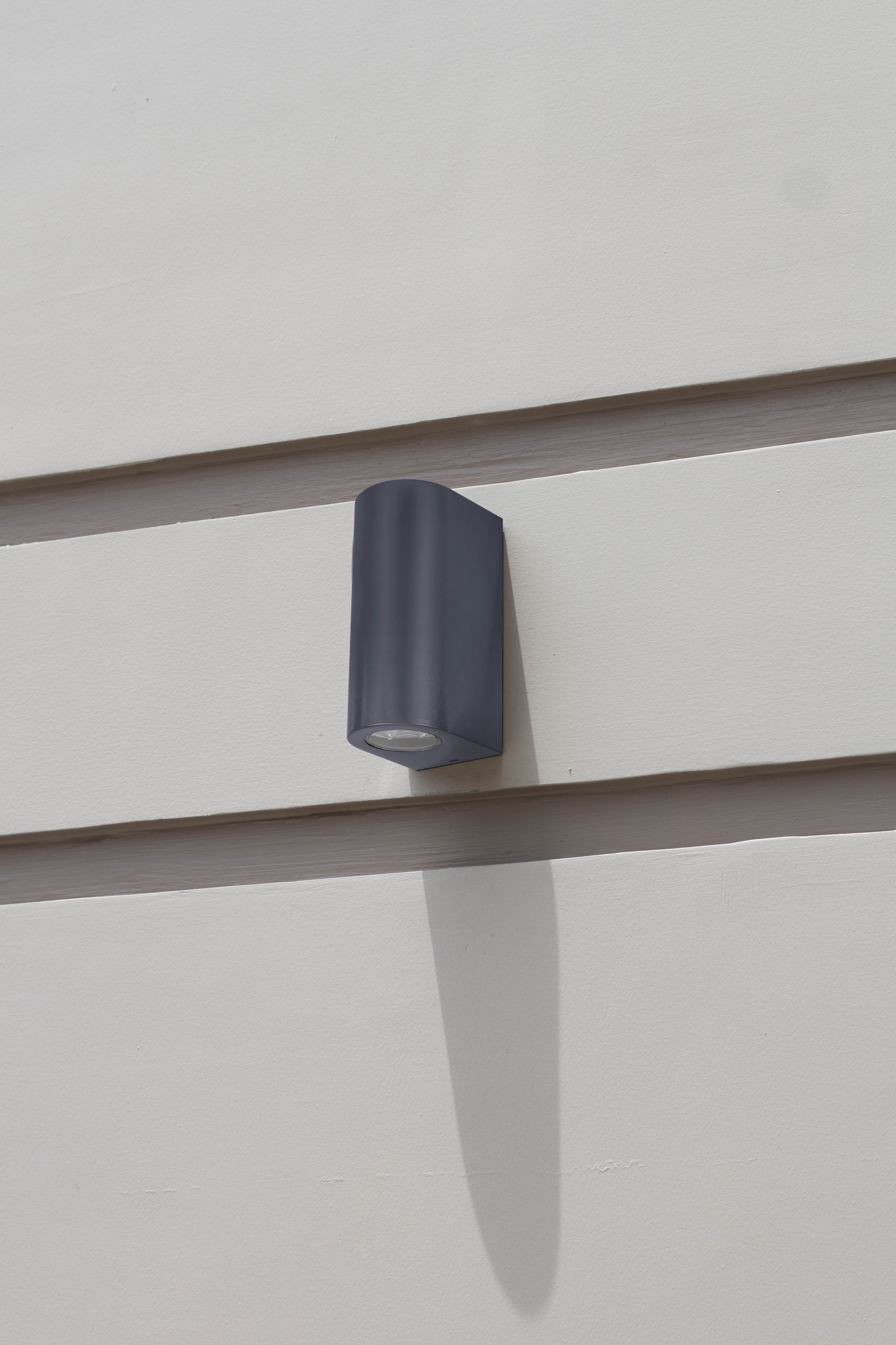 Eine graue Außenlampe an einer weißen Wand. Sie hat ein gebogenes Design und wirft einen Schatten.