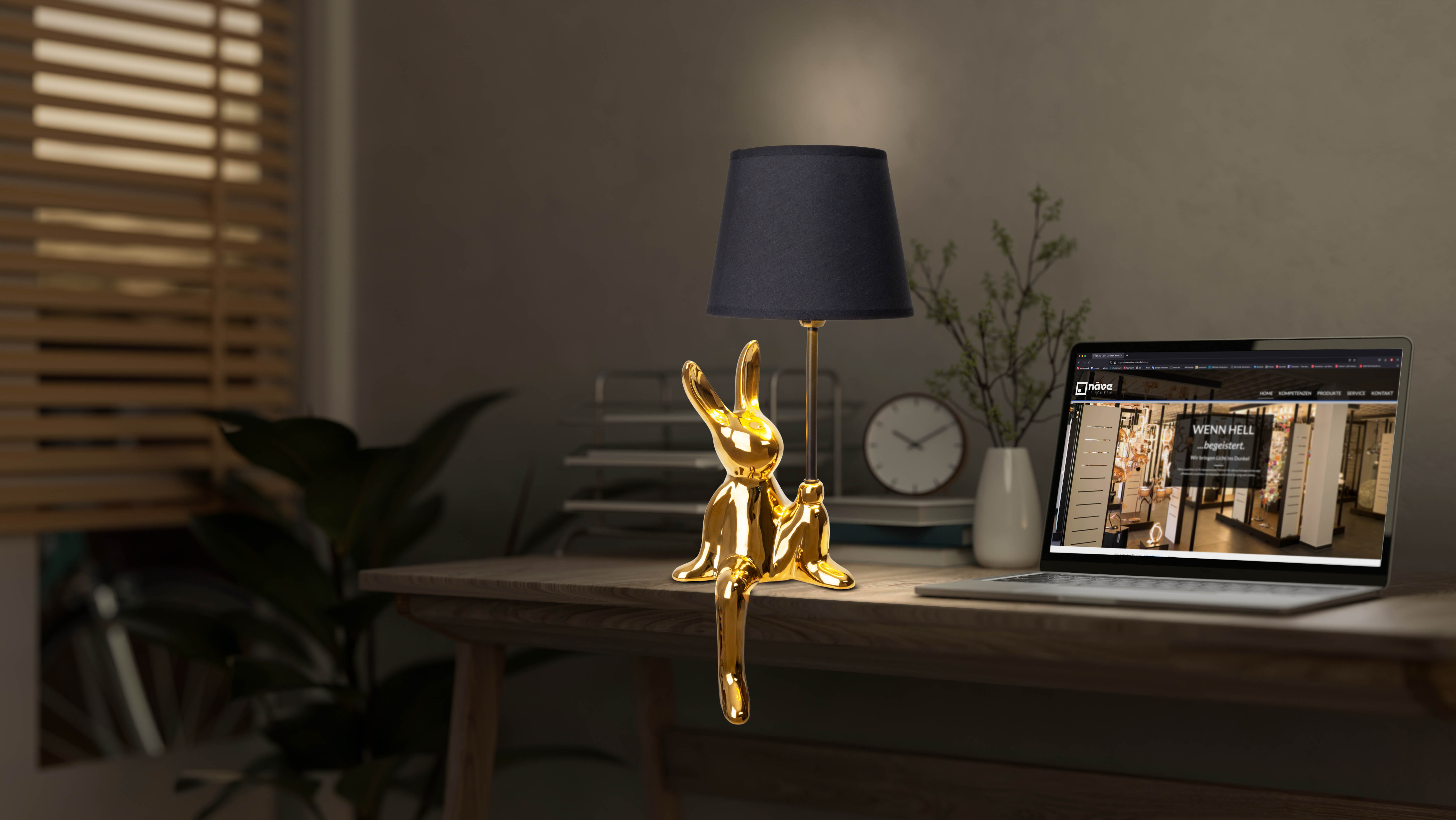 Eine goldene Hasenlampe mit schwarzem Lampenschirm auf einem Holzschreibtisch, neben einem Laptop und einer Uhr.