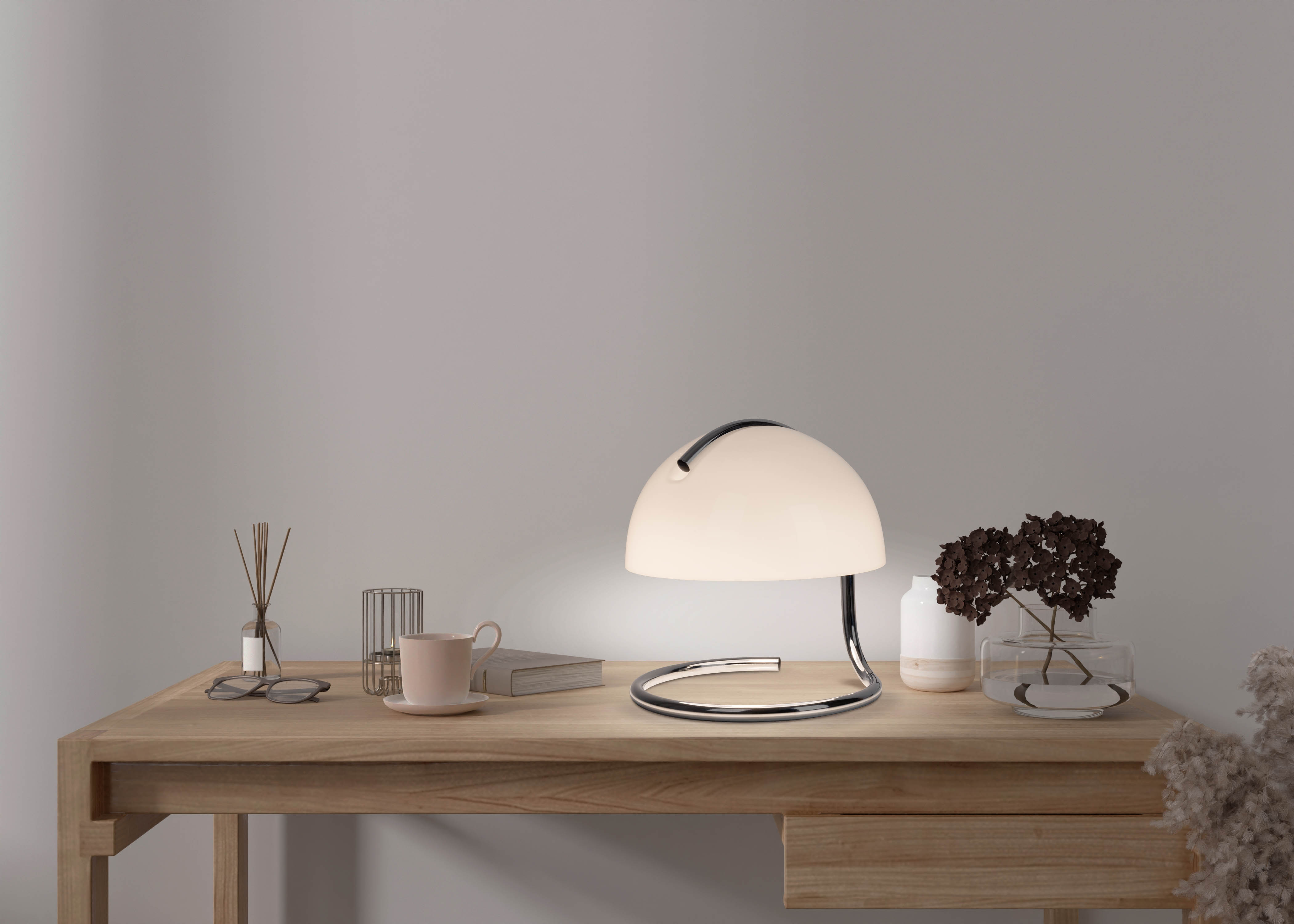 Eine weiße Tischlampe mit geschwungenem Chromfuß auf einem Holzschreibtisch mit Objekten.