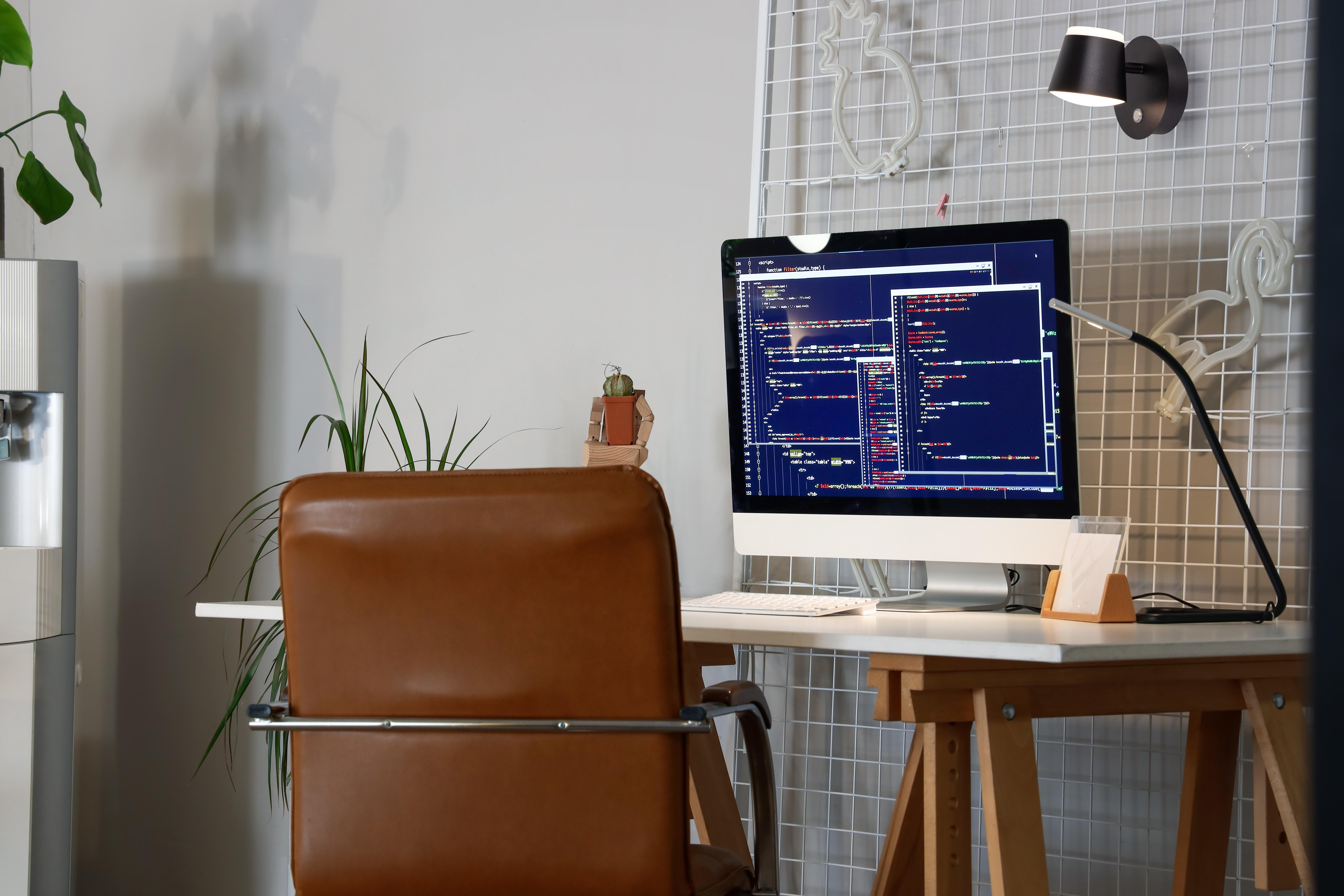 Arbeitsbereich mit Computer, der Code anzeigt, braunem Stuhl, Schreibtisch und Pflanze. Helle Büroumgebung.