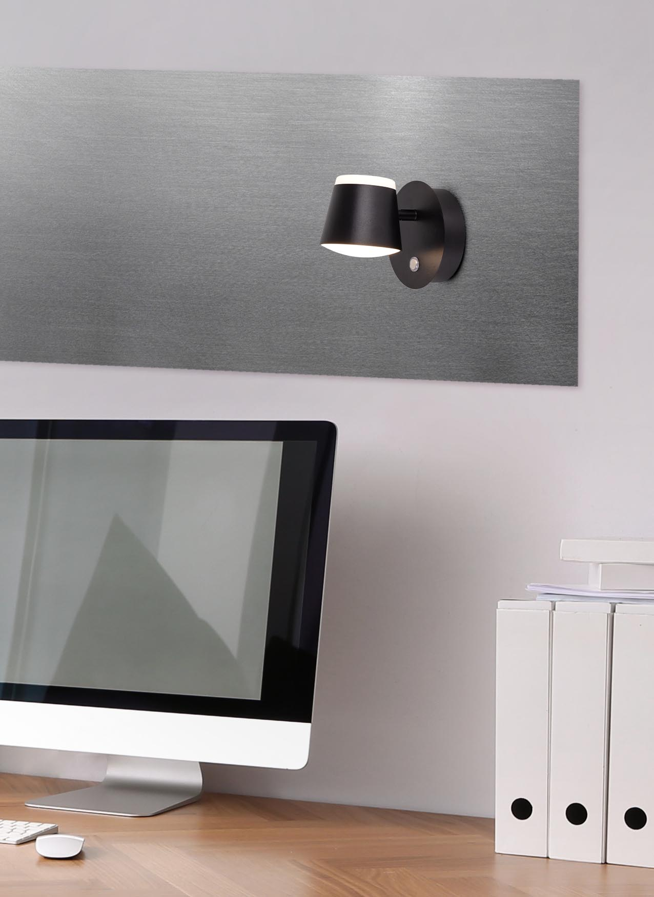 Eine moderne schwarze Lampe an einer grauen Metallwand, mit einem Holzschreibtisch und Computer im Vordergrund.