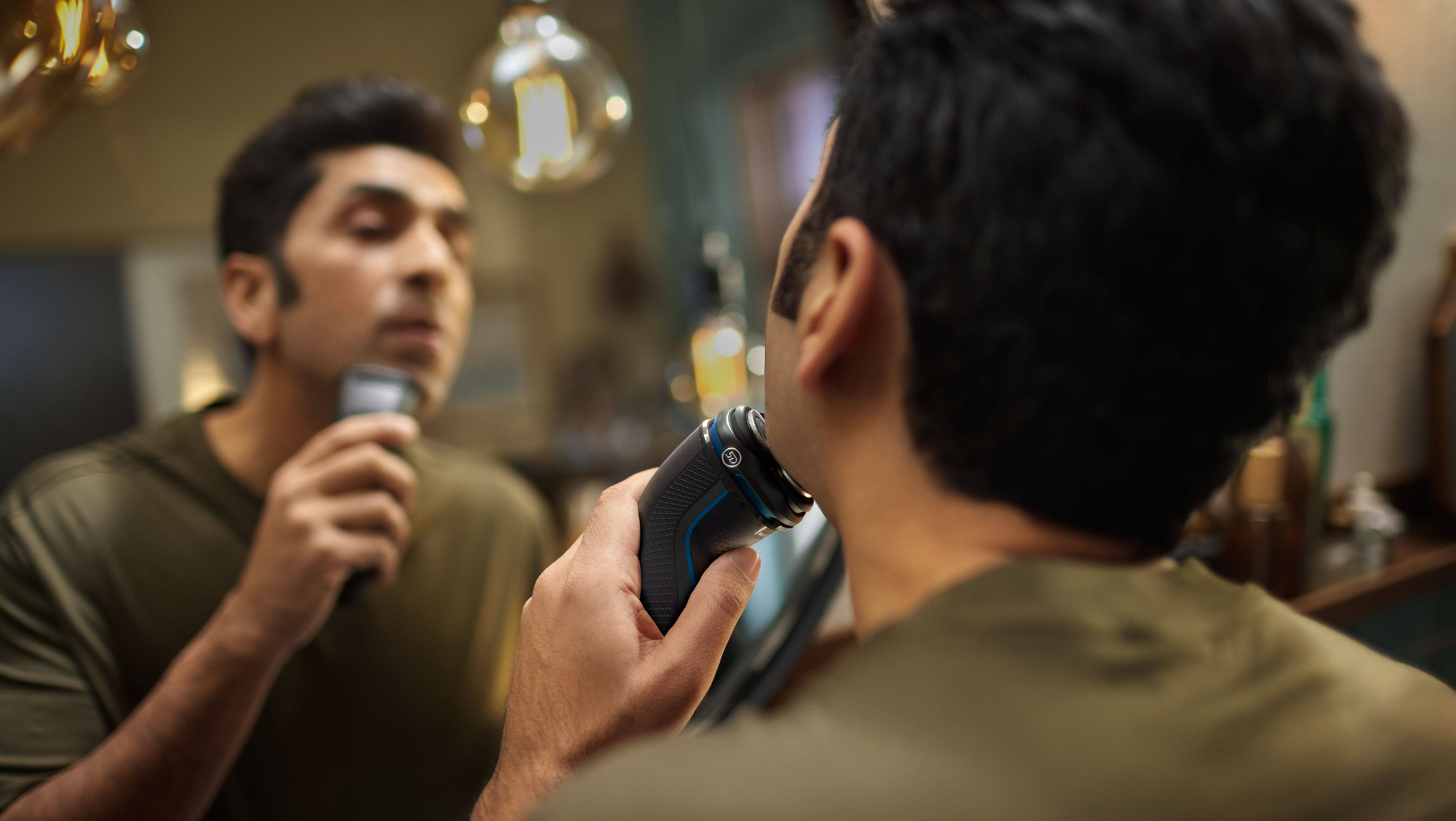 Mężczyzna goli się czarno-niebieską golarką elektryczną przed lustrem.