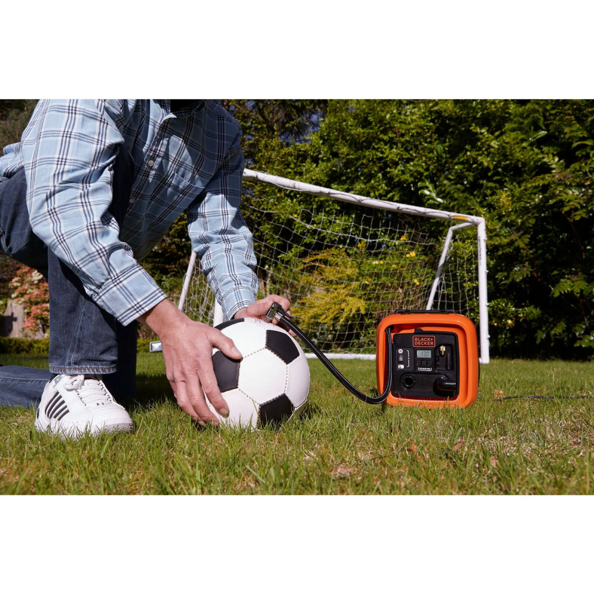 Mann kniend pumpt Fußball mit orangefarbenem Luftkompressor auf Rasen auf, Tor im Hintergrund.