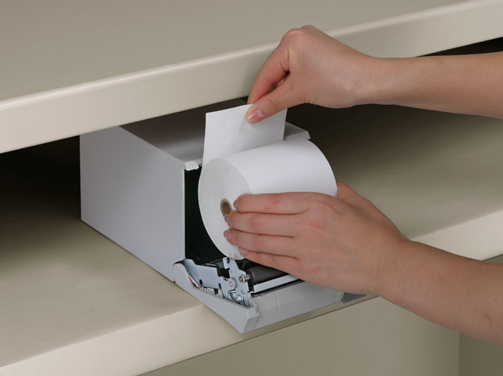 Mains chargeant un rouleau de papier dans une imprimante. L'imprimante est à l'intérieur d'une étagère. Le papier est blanc.