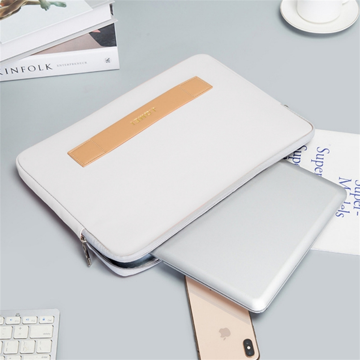 Laptop in weißer Hülle mit braunem Griff, neben Telefon und Büchern. Graue Oberfläche.